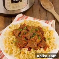 A platter of Hungarian beef goulash on a bed of wide egg noodles, with a wooden spoon and Instant Pot in the background.