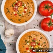 Two bowls of gazpacho soup on a rustic tile background surrounded by some of the ingredients and garnished with cherry tomatoes, cucumber and fresh parsley.