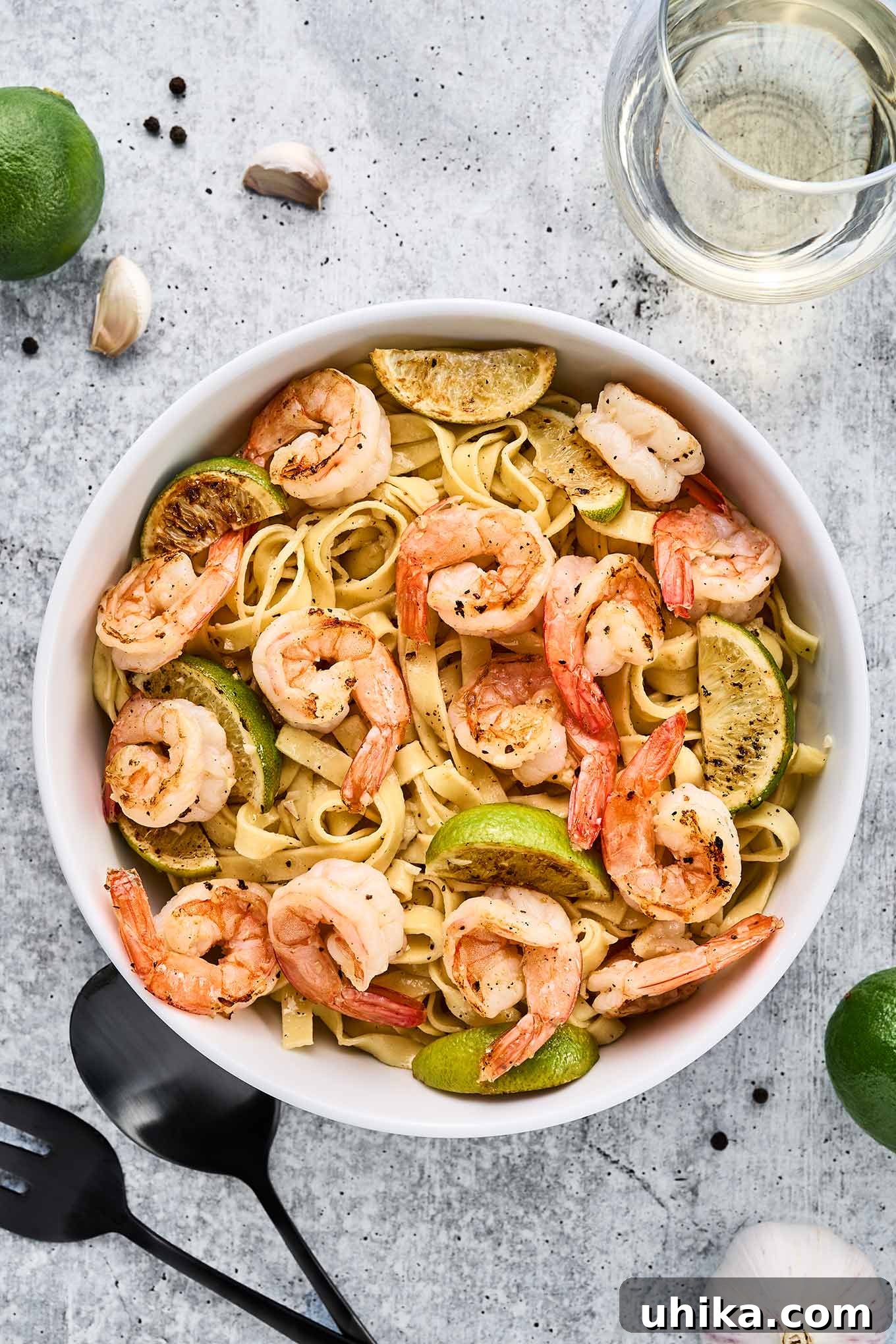 Vibrant Lime Garlic Shrimp Pasta 6 Garlic lime shrimp pasta garnished with lime wedges and fresh herbs in a white bowl on a light stone surface, surrounded by garlic cloves, whole peppercorns, fresh limes, and a bottle of white wine.