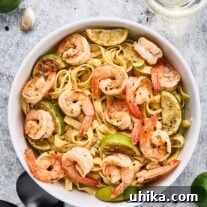 Garlic lime shrimp pasta garnished with lime wedges in a white bowl on a light stone surface, surrounded by garlic cloves, peppercorns, limes, and white wine.