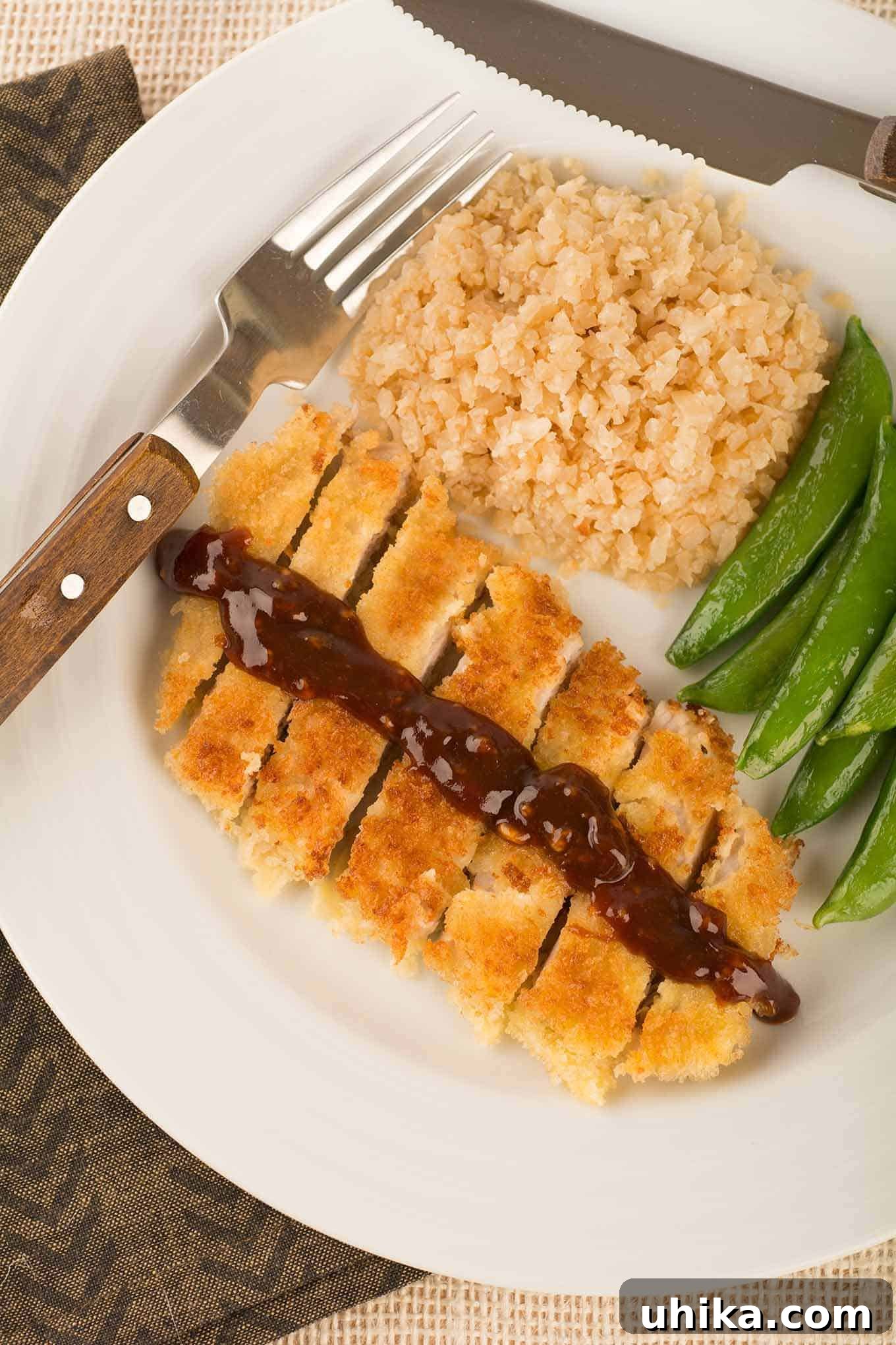 Crispy Japanese Tonkatsu 2 An overhead view of a plate with Japanese breaded pork cutlet, sliced and topped with a drizzle of katsu sauce, served with riced cauliflower and sautéed snap peas. This classic dish features crispy panko-coated pork.