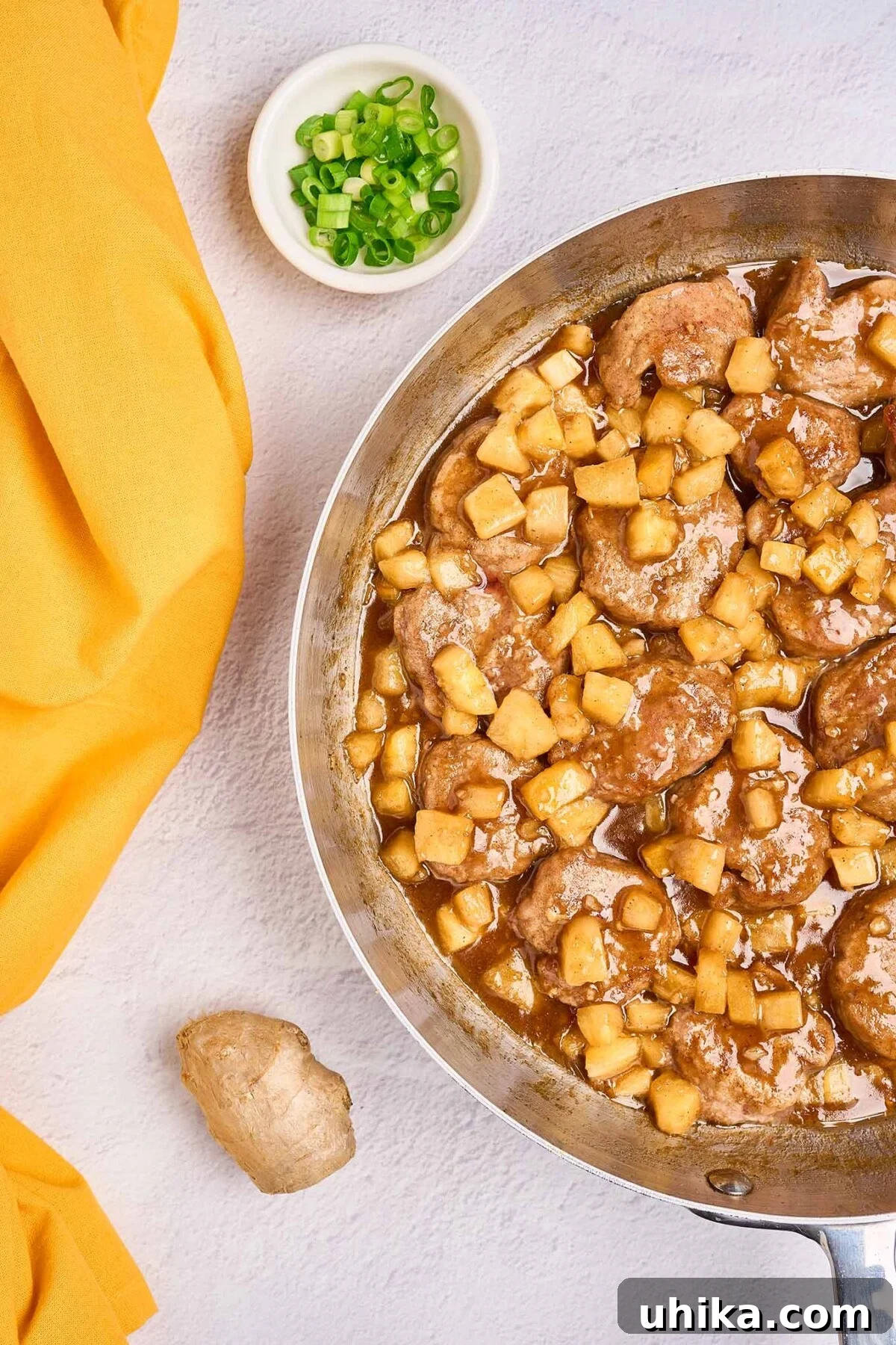 Island-Style Pineapple Pork Skillet 4 A skillet filled with tender pork medallions and pineapple chunks swimming in a sweet-savory rum sauce, with a small bowl of chopped scallions and a knob of fresh ginger to the side, showcasing the preparation process.