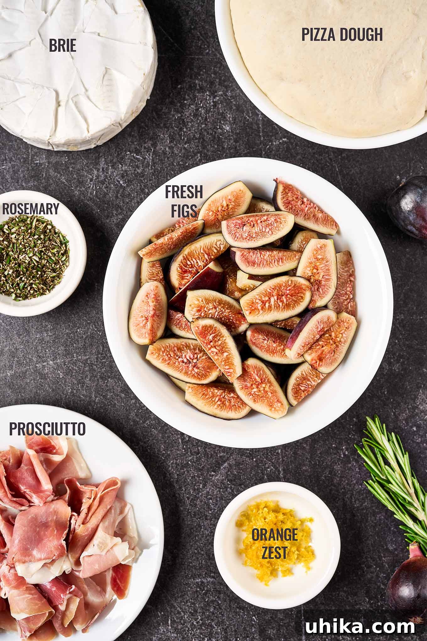 Gourmet Fig Prosciutto Brie Pizza 3 A labeled, overhead view of the ingredients for fig and prosciutto pizza including fresh figs, pizza dough, prosciutto, rosemary, orange zest, and brie, beautifully arranged on a rustic board.