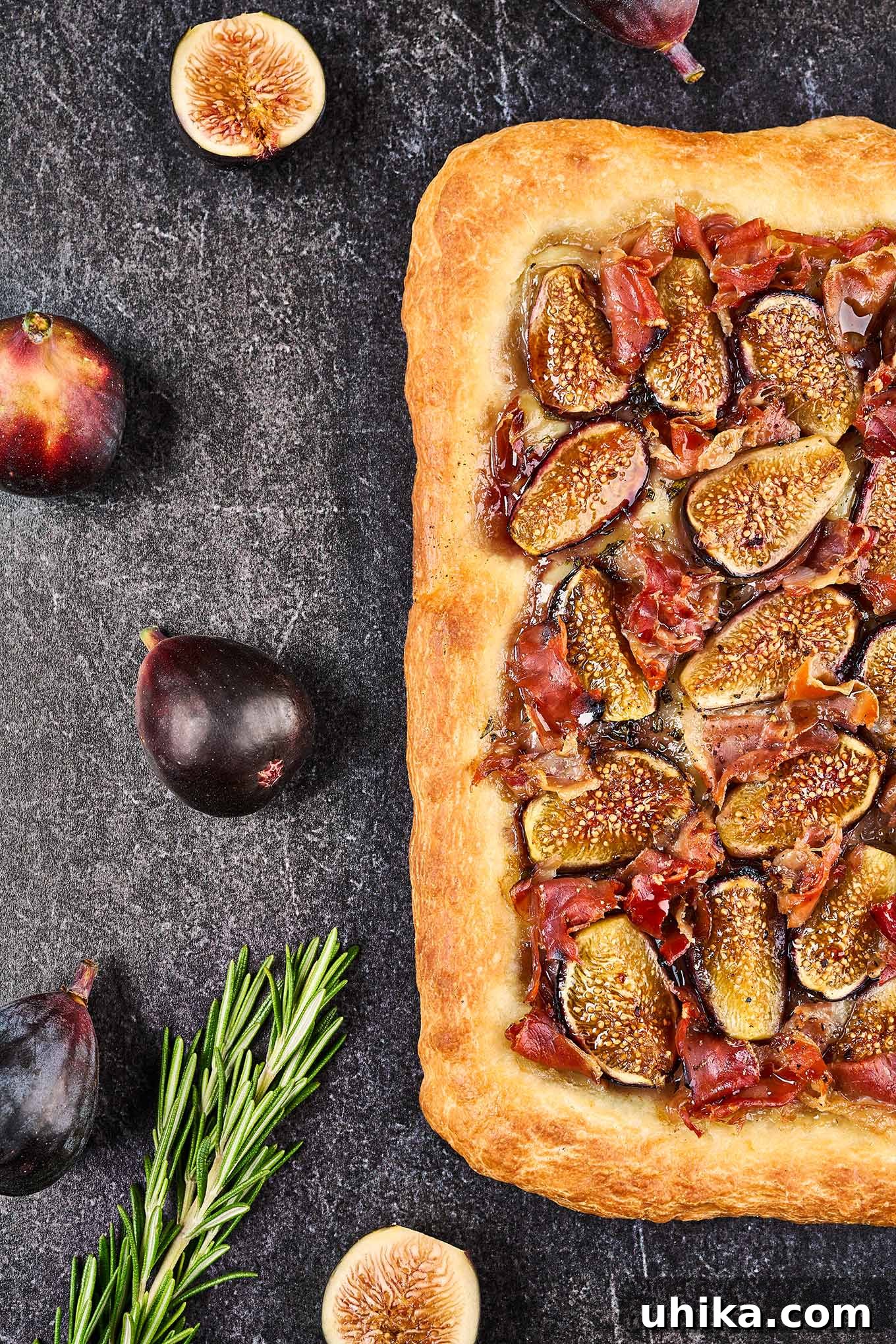 Gourmet Fig Prosciutto Brie Pizza 5 A freshly baked fig and prosciutto pizza on a slate background next to whole and cut figs and sprigs of fresh rosemary, highlighting its gourmet appeal.