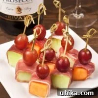 Close-up of a prosciutto-wrapped melon bite, garnished with a red grape and secured with a small wooden skewer, ready for serving.