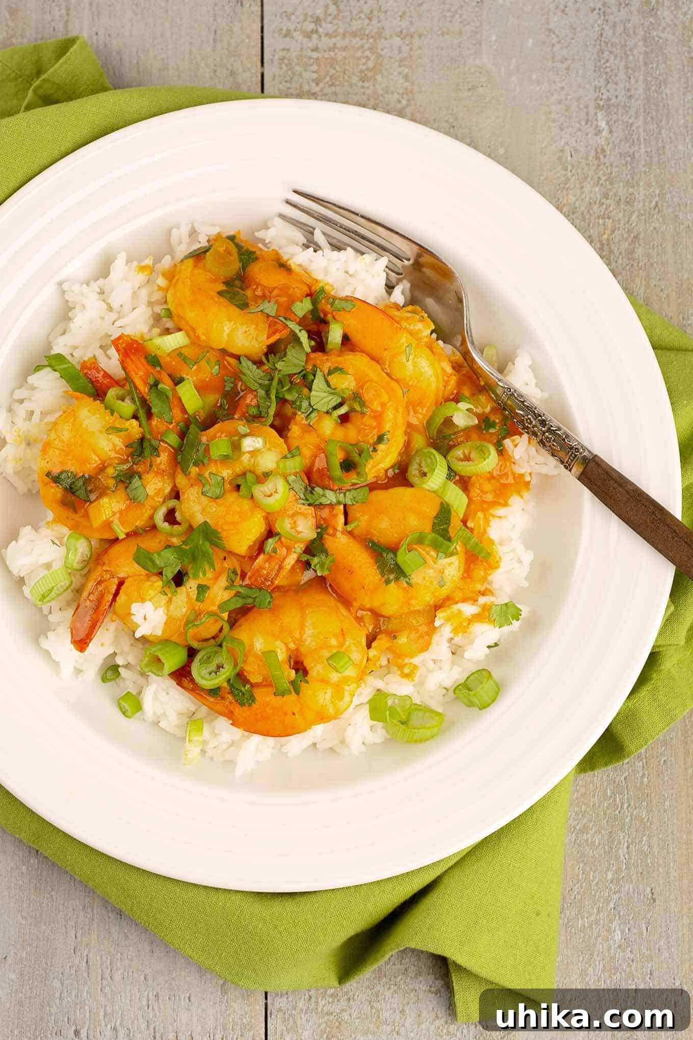 Creamy Coconut Curry Shrimp 2 An overhead view of a bowl of coconut curry shrimp over rice, scallion and cilantro garnish, a fork, and a napkin underneath the bowl.