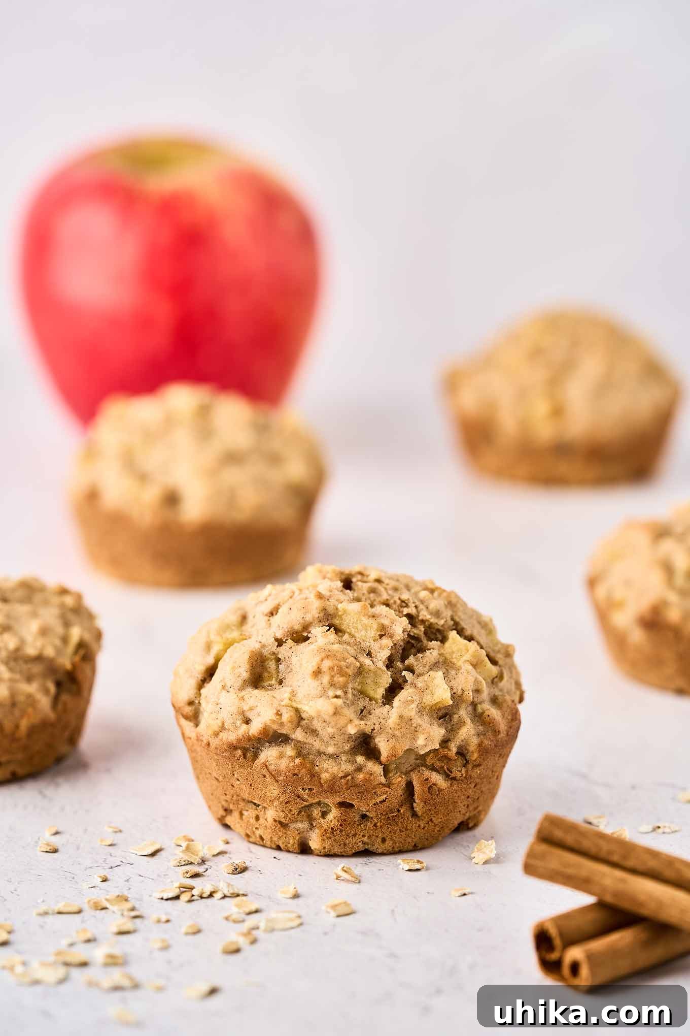 Cozy Apple Cinnamon Oat Muffins 2 A single apple oatmeal muffin with more muffins and a whole apple in the background, cinnamon sticks, and scattered oats in the foreground. This wholesome muffin offers a delicious start to any day.