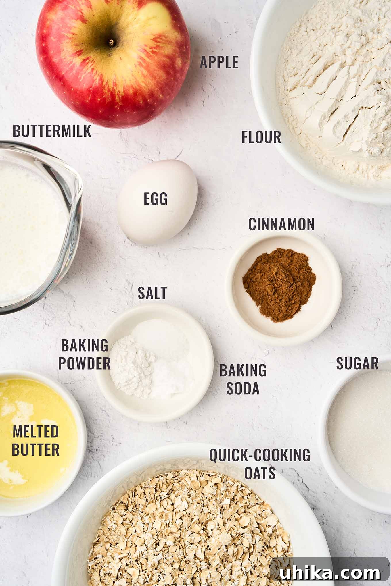 Cozy Apple Cinnamon Oat Muffins 3 A labeled, overhead view of the ingredients needed to make apple cinnamon oatmeal muffins; including flour, oats, sugar, cinnamon, egg, melted butter, buttermilk, apple, baking soda, baking powder, and salt.