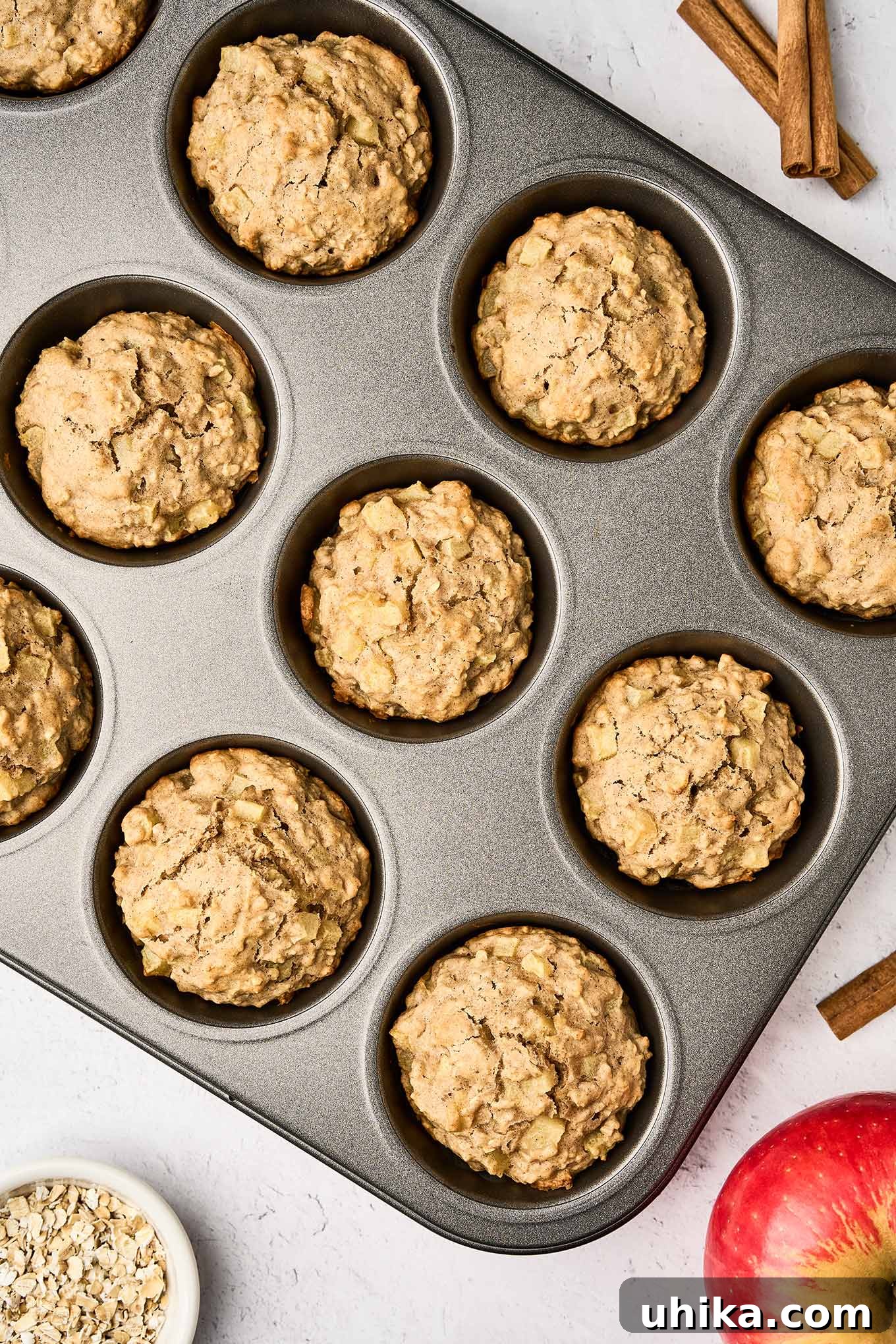 Cozy Apple Cinnamon Oat Muffins 6 An overhead view of warm apple oatmeal muffins fresh from the oven in their muffin tin, elegantly arranged with a whole apple, cinnamon sticks, and a small bowl of oats, embodying the essence of homemade comfort.