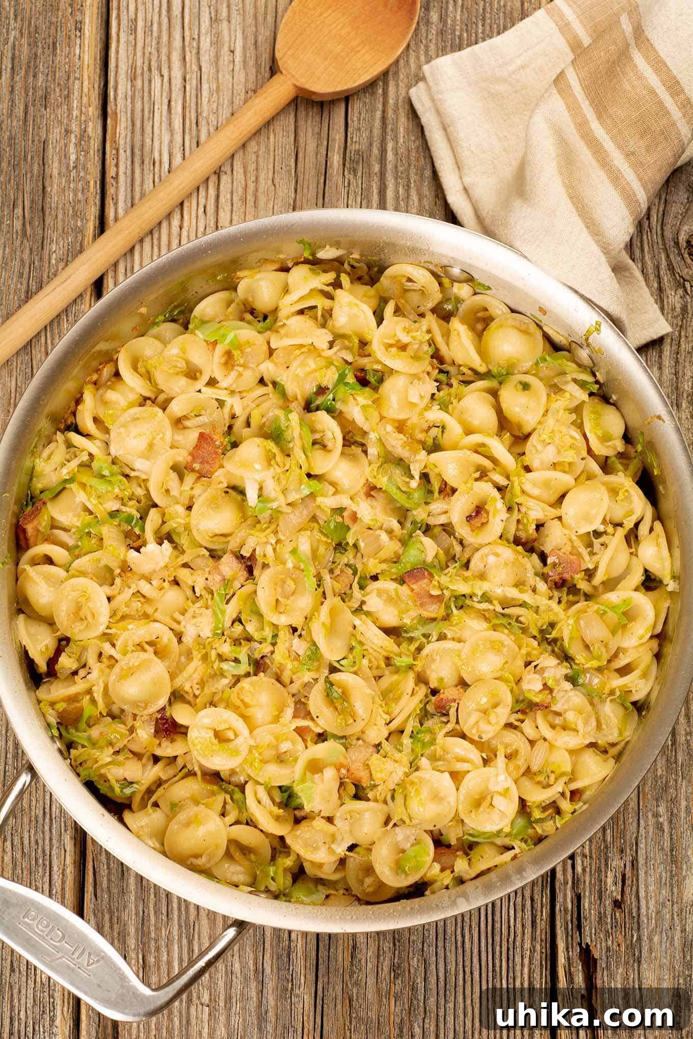 Golden Orecchiette with Smoky Bacon and Roasted Brussels 2 A sizzling sauté pan filled with orecchiette pasta, perfectly tossed with golden-brown shredded Brussels sprouts, crispy bacon pieces, caramelized onions, and fragrant garlic. A rustic wooden spoon is poised in the background, alongside a kitchen towel, hinting at the culinary effort and inviting taste.