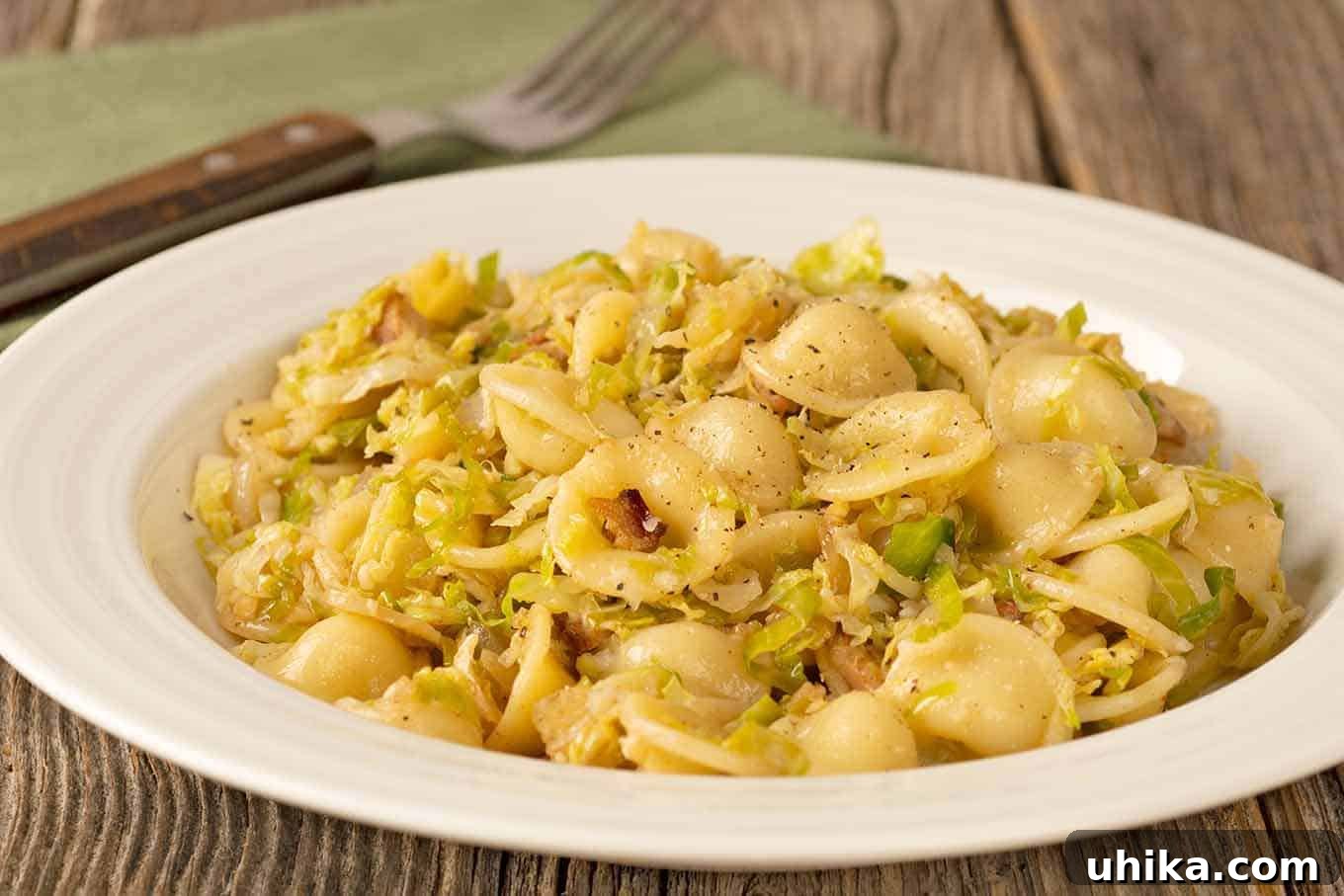 Golden Orecchiette with Smoky Bacon and Roasted Brussels 3 A mouth-watering close-up of a serving of orecchiette pasta mingled with tender, finely shredded Brussels sprouts and generous pieces of golden, crispy bacon on a white plate. A fork is placed casually next to the dish, with a soft, textured napkin subtly in the background, showcasing the inviting simplicity and delectable appeal of the meal.