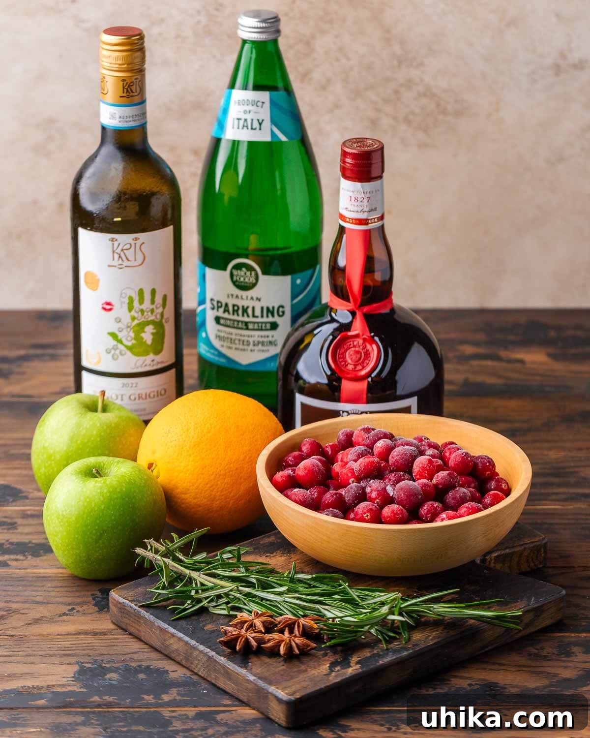 Spiced Christmas Sangria 3 All the essential ingredients for Christmas Sangria laid out: a bottle of dry white wine, sparkling water, a bottle of Grand Marnier, fresh green apples, a vibrant orange, a bowl of red cranberries, fresh rosemary sprigs, and fragrant star anise pods.