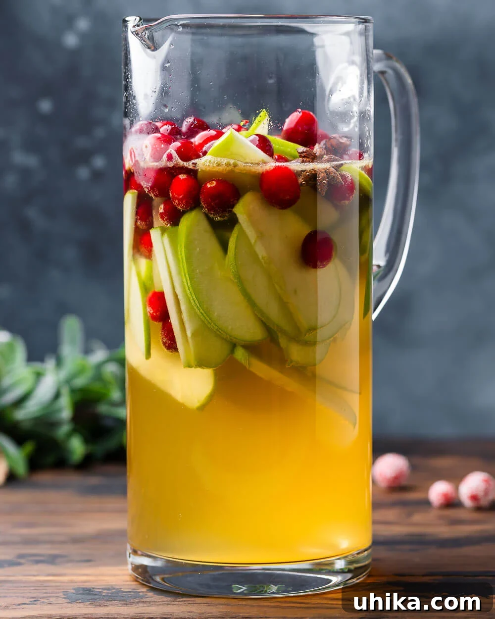 Spiced Christmas Sangria 4 A large pitcher filled with vibrant Christmas Sangria, brimming with cranberries and apple slices, beautifully garnished with rosemary and star anise, set against a subtle blue background.