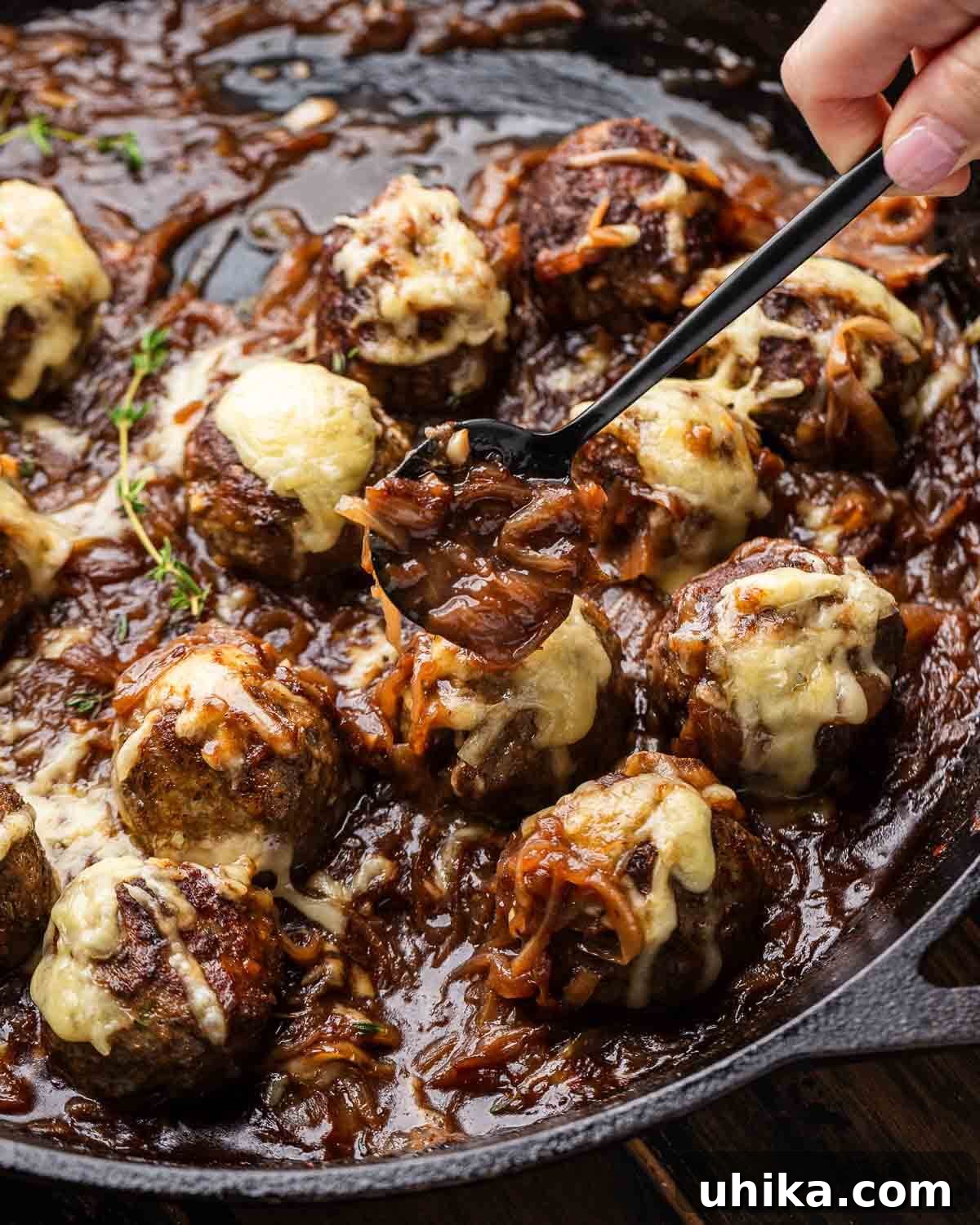 Bistro French Onion Meatballs 2 Hands holding spoonful of golden caramelized onions over perfectly browned French onion meatballs in a rustic cast iron pan, ready to be served.
