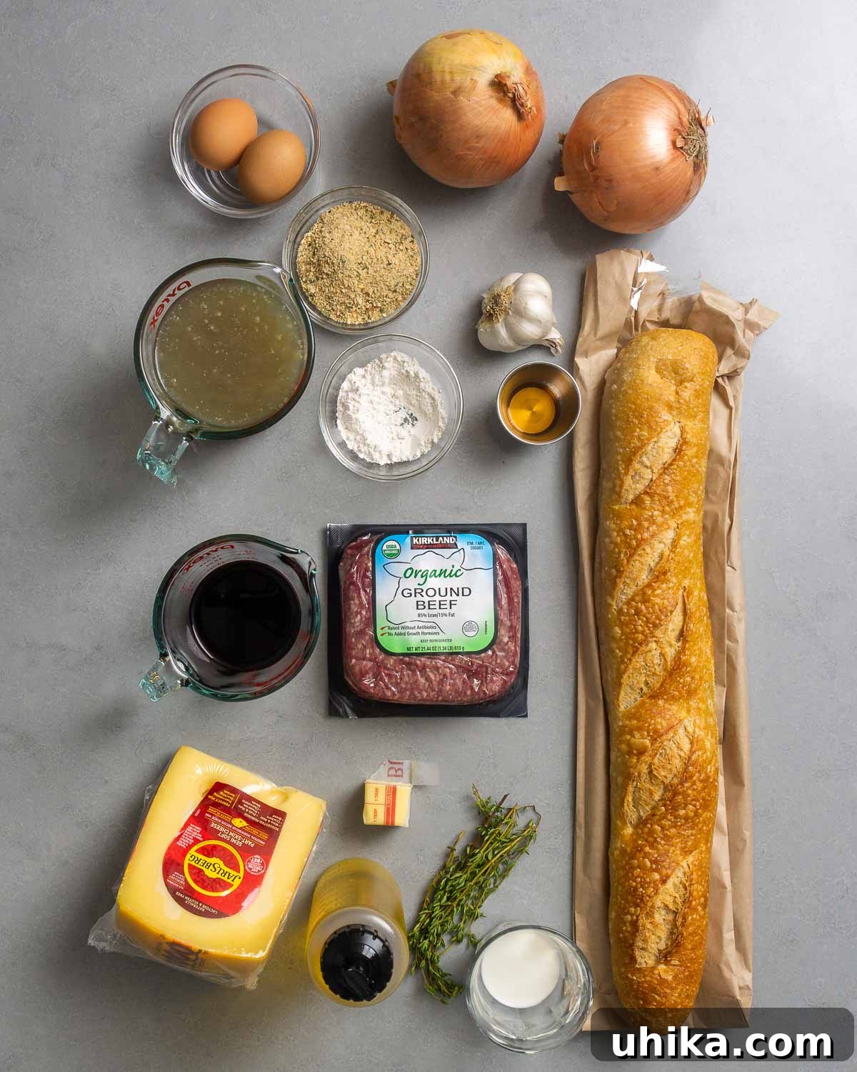 Bistro French Onion Meatballs 3 A comprehensive flat lay of all ingredients for French onion meatballs: eggs, chicken stock, breadcrumbs, flour, yellow onions, fresh garlic, brandy, baguette slices, red wine, ground beef, Swiss cheese, butter, olive oil, fresh thyme, and milk, all arranged on a rustic wooden surface.