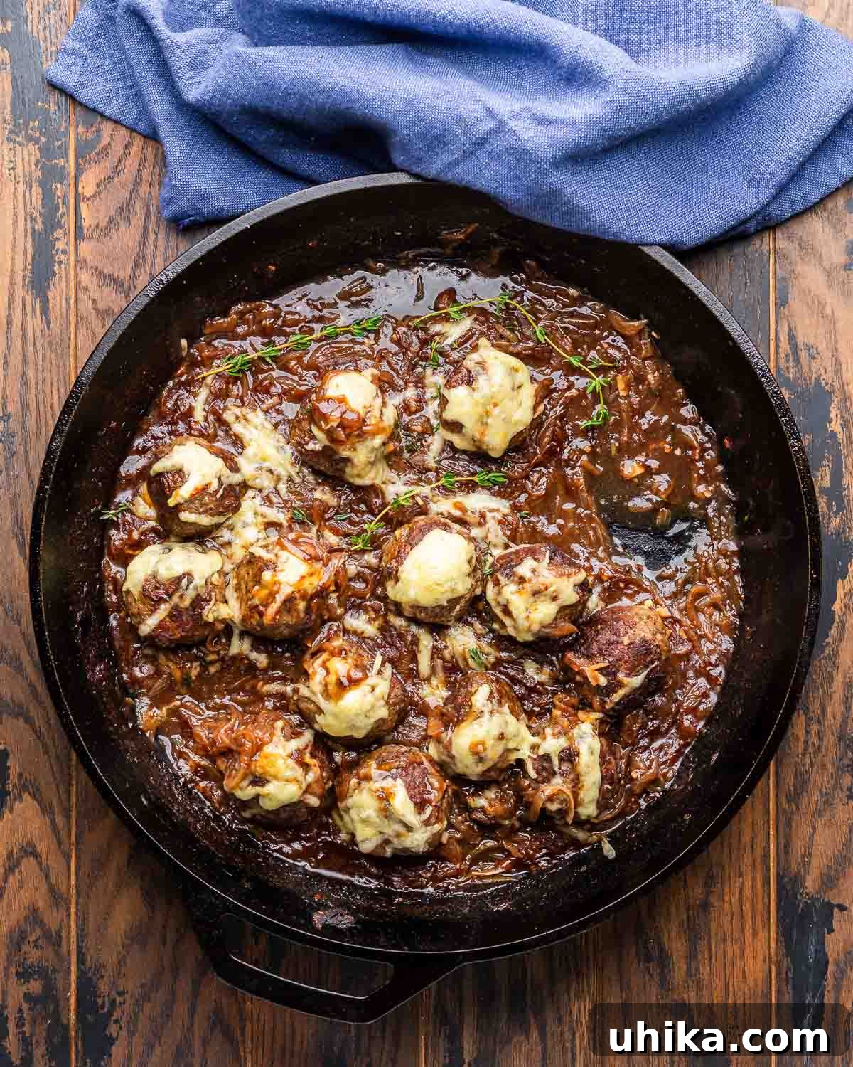 Bistro French Onion Meatballs 8 Close-up of French onion meatballs simmering in a rich, bubbling caramelized onion and red wine sauce, topped with melted Swiss cheese in a cast iron pan, with a blue kitchen towel draped nearby.