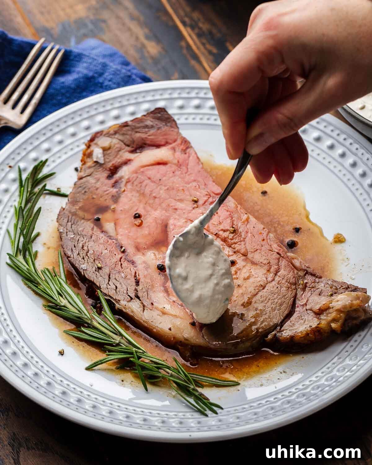 Tender Prime Rib with Rich Beef Drippings 8 Hands spooning horseradish sauce onto a slice of prime rib in white plate.