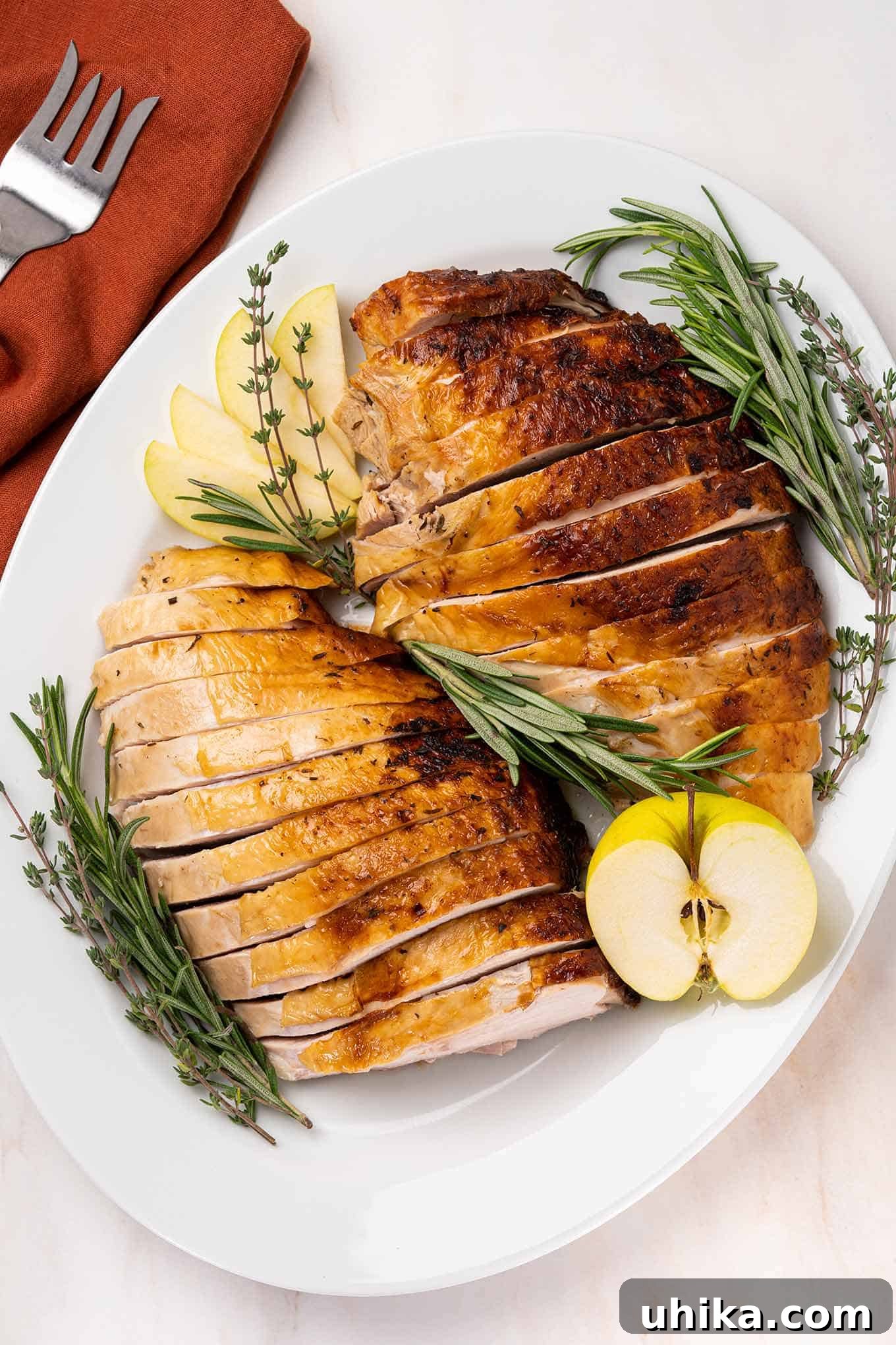 Sweet & Savory Apple Cider Turkey Breast 2 An overhead view of a carved turkey breast with golden brown skin on an oval serving platter garnished with fresh herbs and apple.
