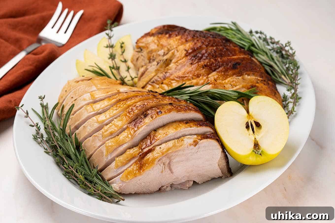 Sweet & Savory Apple Cider Turkey Breast 7 Carved turkey breast on a platter garnished with fresh rosemary and thyme and half an apple, serving fork in the background.