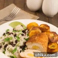 A dinner plate with a crumb-coated, rolled boneless chicken breast stuffed with sweet plantains, served with black beans and rice and sautéed maduros, a fork, napkin, and salt and pepper shakers in the background.