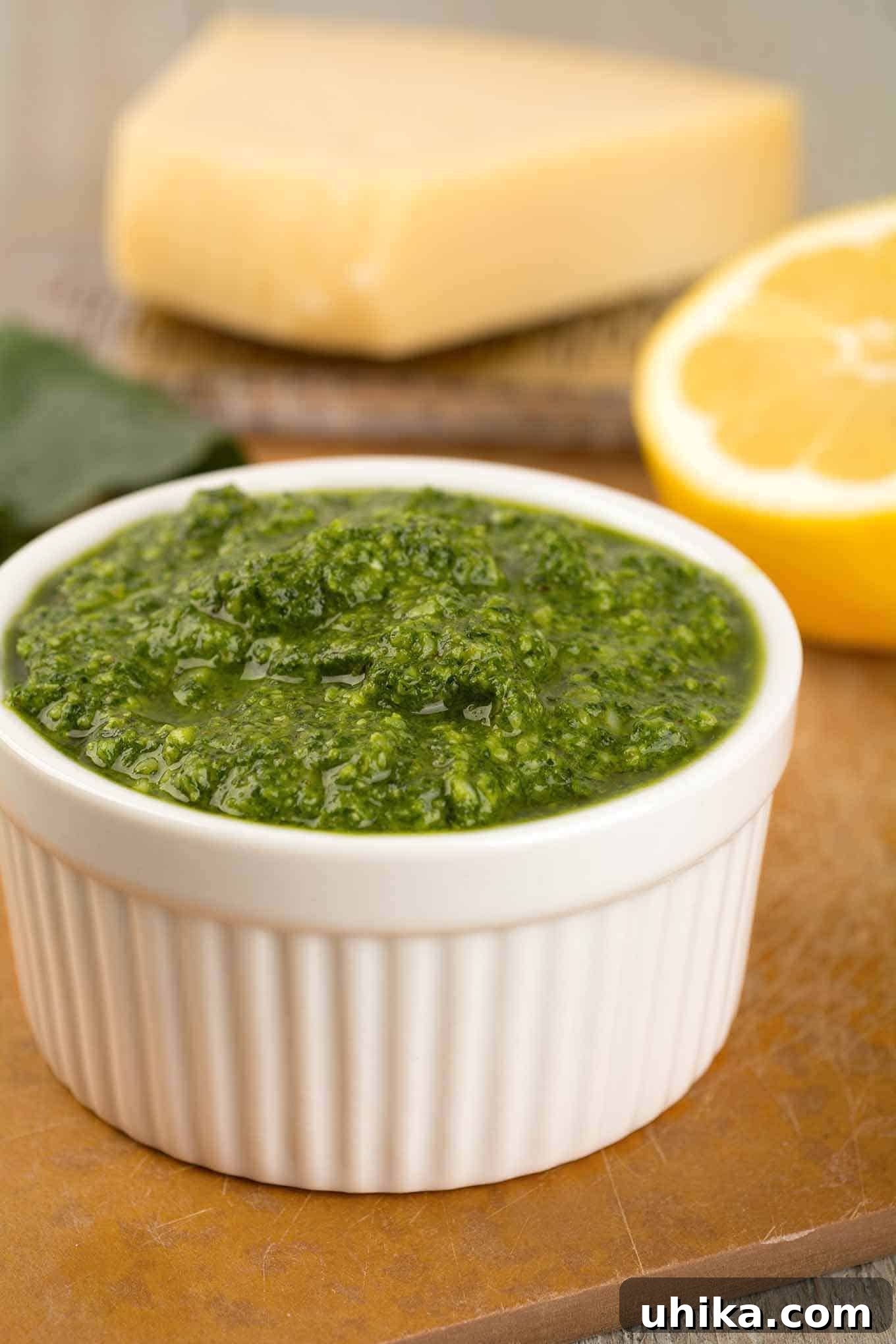 Vibrant Kale Pesto 2 A small white bowl filled with vibrant green kale pesto, accompanied by a halved lemon and a wooden cutting board featuring a wedge of Parmesan cheese in the background. This image highlights the fresh ingredients and appetizing appearance of homemade kale pesto.