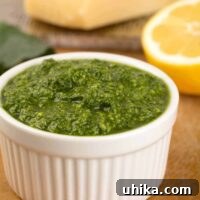 A small white bowl filled with vibrant green kale pesto, accompanied by a halved lemon and a wooden cutting board featuring a wedge of Parmesan cheese in the background. This image highlights the fresh ingredients and appetizing appearance of homemade kale pesto.