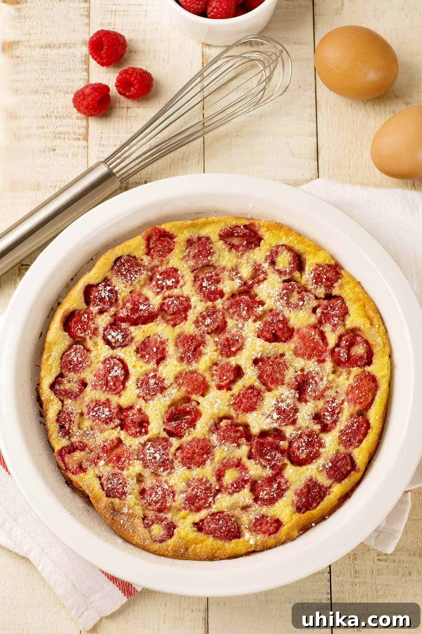 Berry-Kissed Custard Delight 2 An overhead view of freshly baked raspberry clafoutis dusted with powdered sugar in a white pie plate, surrounded by a whisk, eggs, and fresh raspberries. This classic French dessert is simple yet elegant, perfect for any occasion.