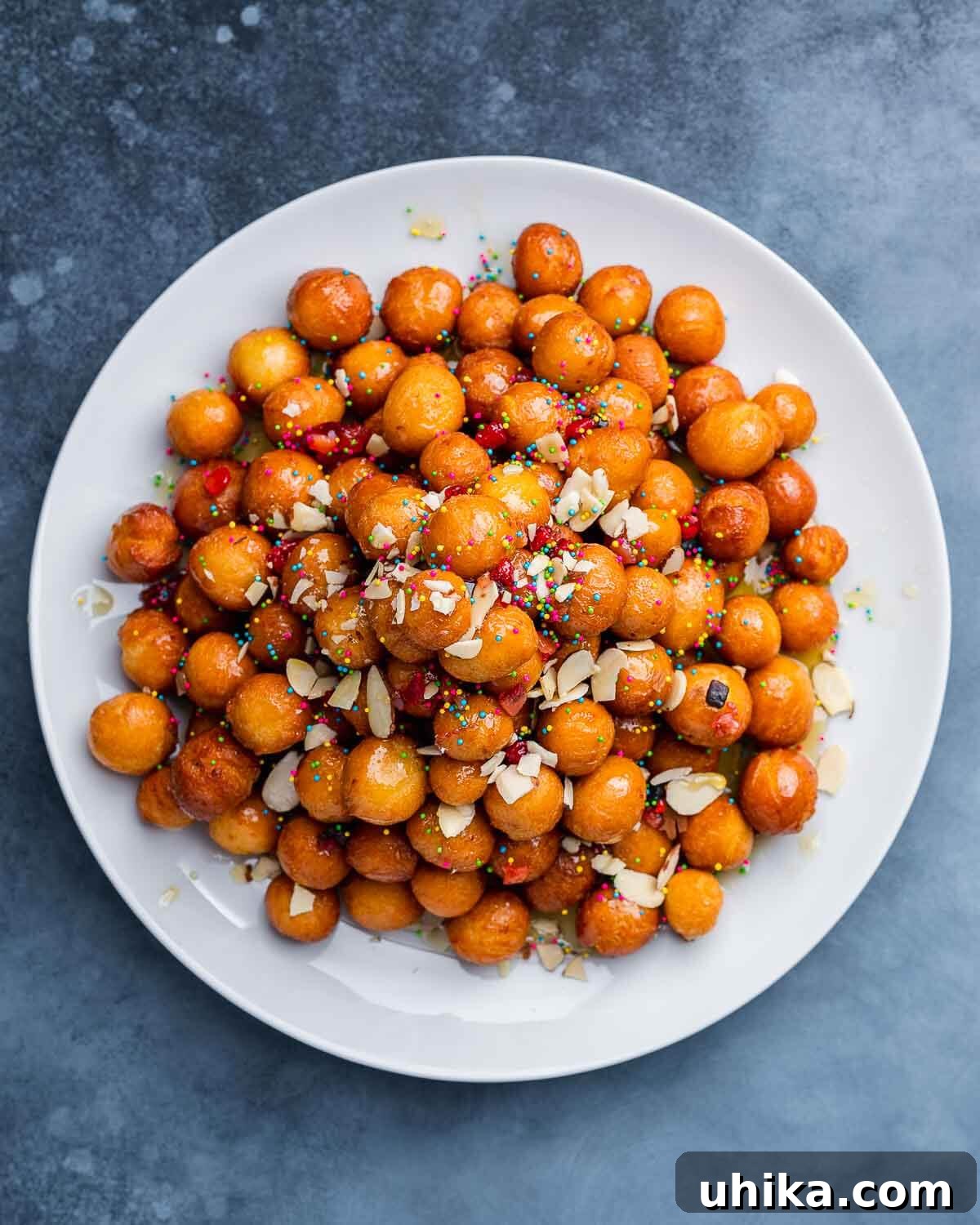 Podcast 27: Christmas Cookies - Must-Bakes and Must-Skips 2 Overhead shot of white platter with golden brown struffoli drizzled with honey and colorful sprinkles on a blue wooden board, perfect for Christmas celebrations.