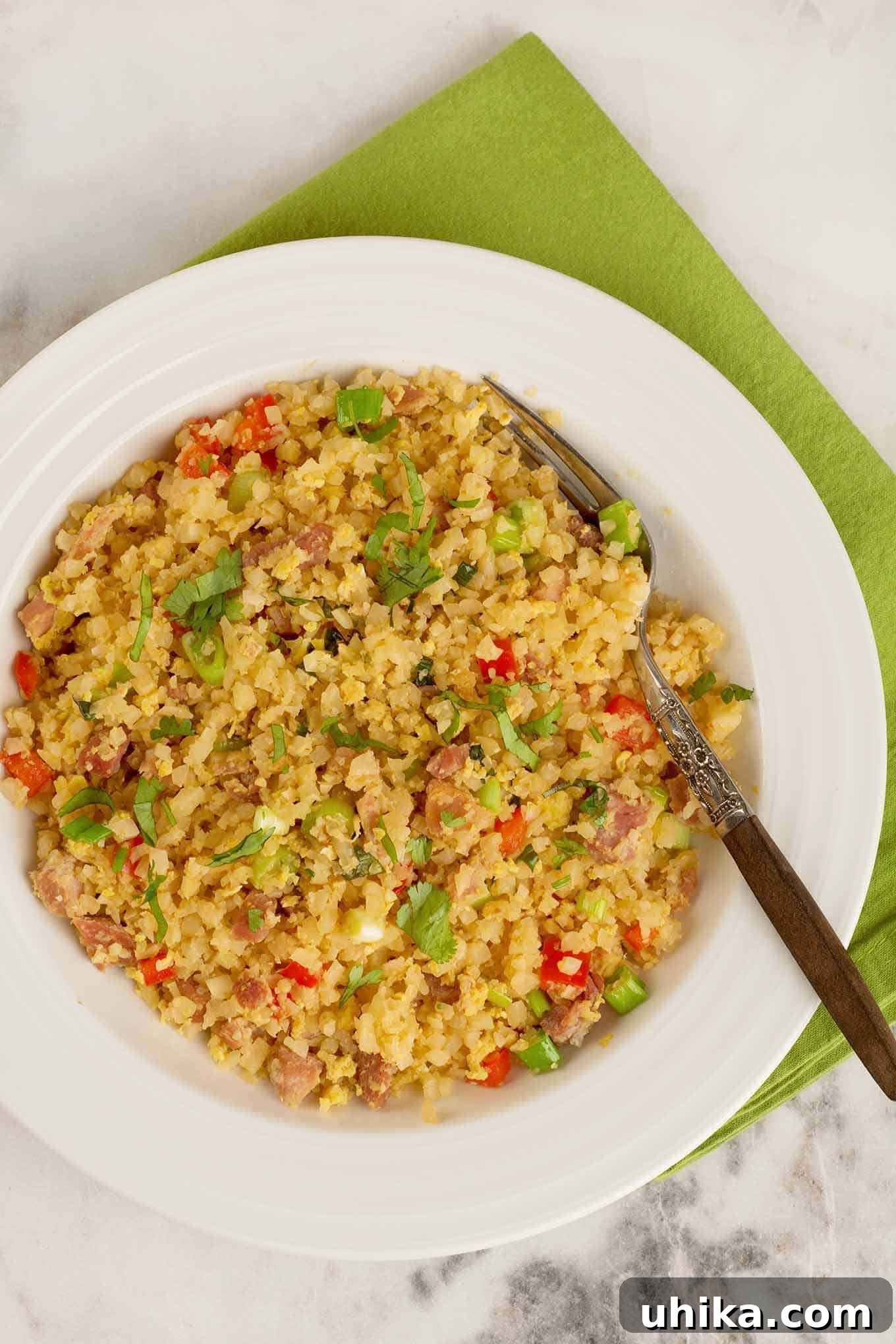 Light Cauliflower Fried Rice 2 An overhead view of a bowl of stir fried riced cauliflower combined with red bell pepper, scallions, scrambled egg, ginger, garlic, and cilantro, a wooden-handled fork to the right.