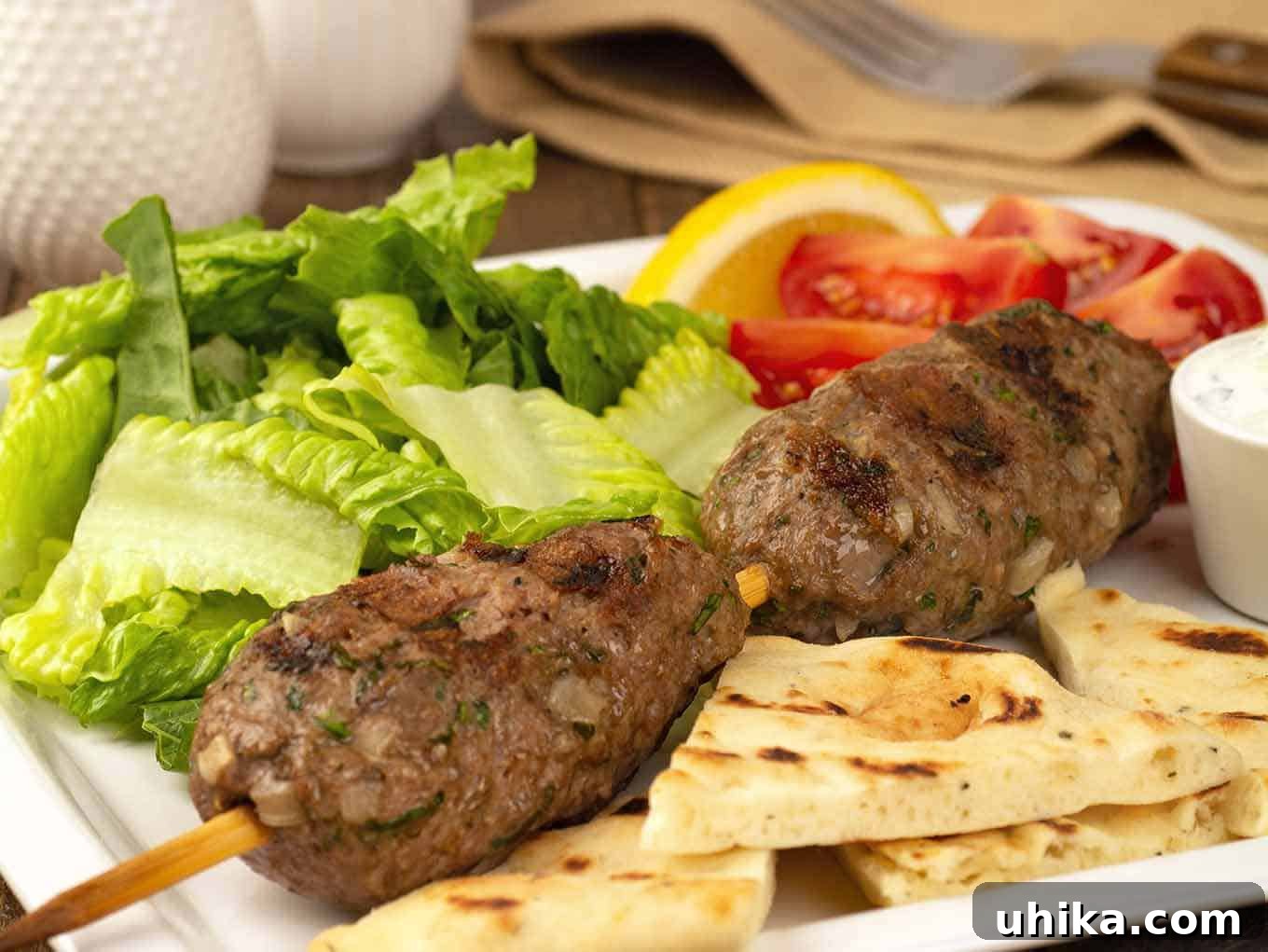 Flame-Grilled Moroccan Lamb Kefta 3 A close view of two grilled, skewered lamb meatballs, pita bread, lettuce, tomatoes, and lemon wedges on a dinner plate.