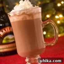 A clear Irish coffee mug filled with rich, creamy Irish hot chocolate, generously topped with fluffy whipped cream and a sprinkle of cocoa powder. Soft, festive Christmas lights glow warmly in the blurred background, creating a cozy and inviting atmosphere perfect for the holidays.