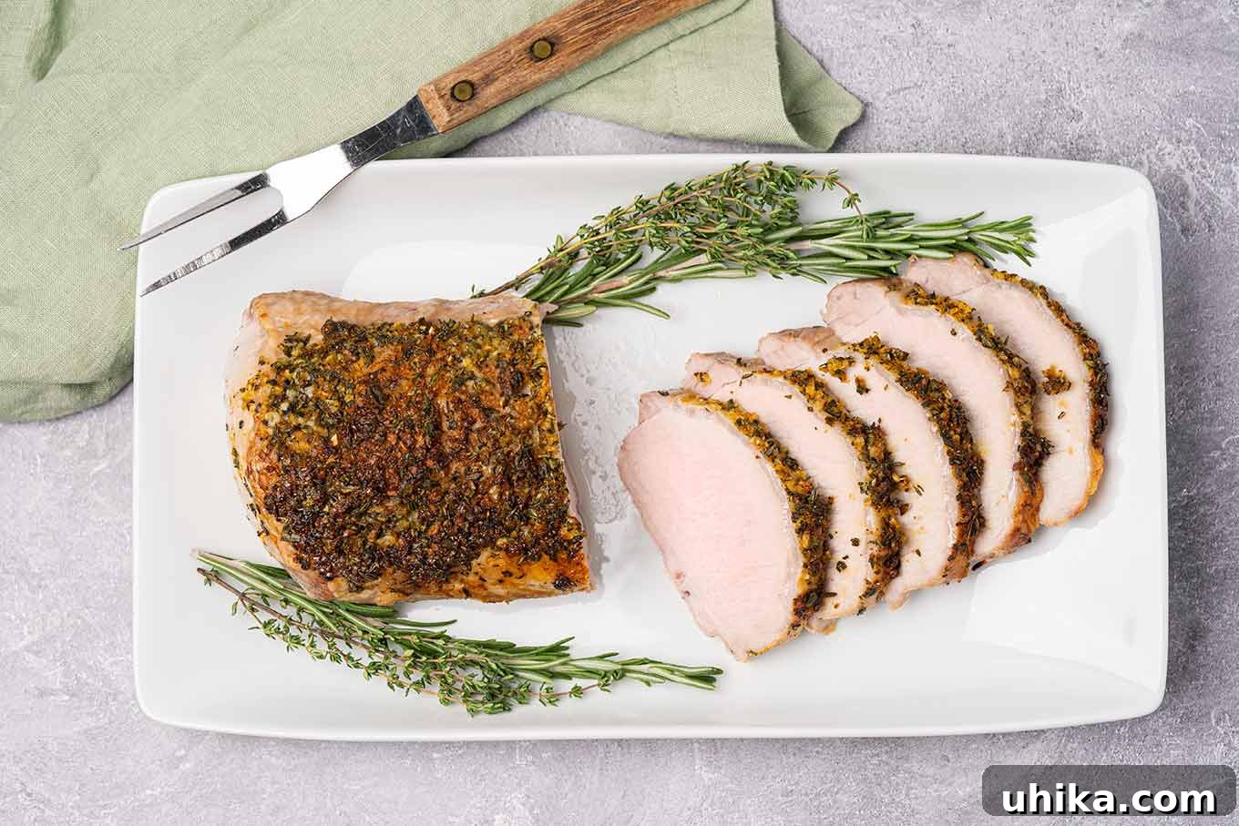 Golden Herb Crusted Pork Loin 5 A partially sliced boneless pork roast on a serving platter with a carving fork, garnished with sprigs of fresh rosemary and thyme.