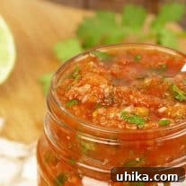 An open jar of homemade salsa, garnished with chopped cilantro, out of focus ingredients on a cutting board in the background.
