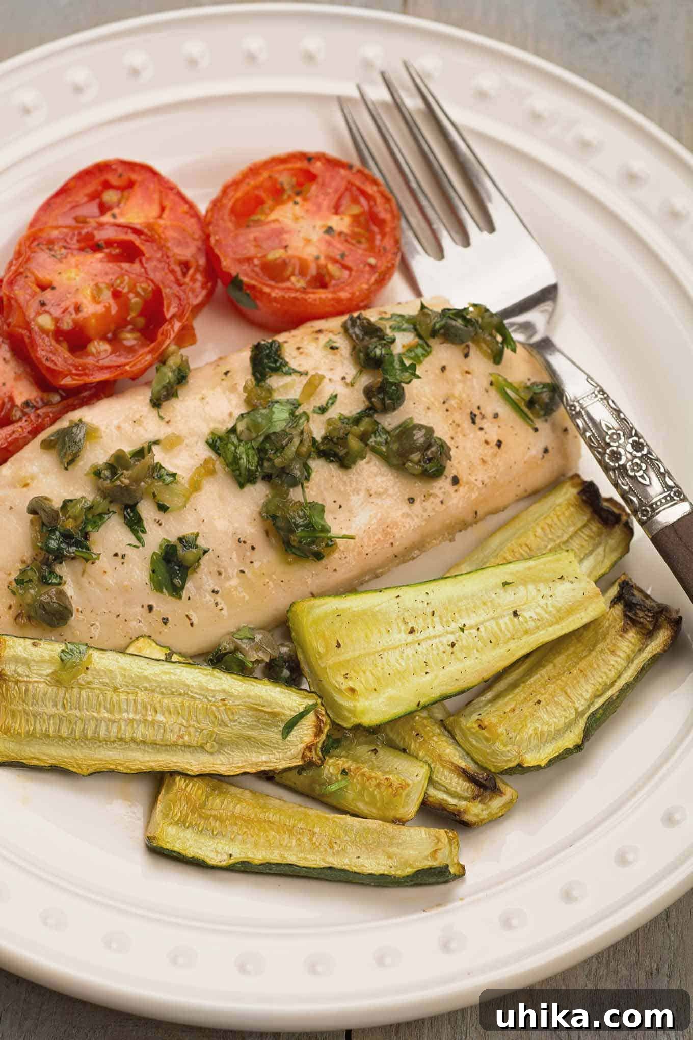 Zesty Mediterranean Traybake Fish 3 A dinner plate with a single tilapia fillet topped with capers and parsley, roasted plum tomato slices and zucchini wedges.