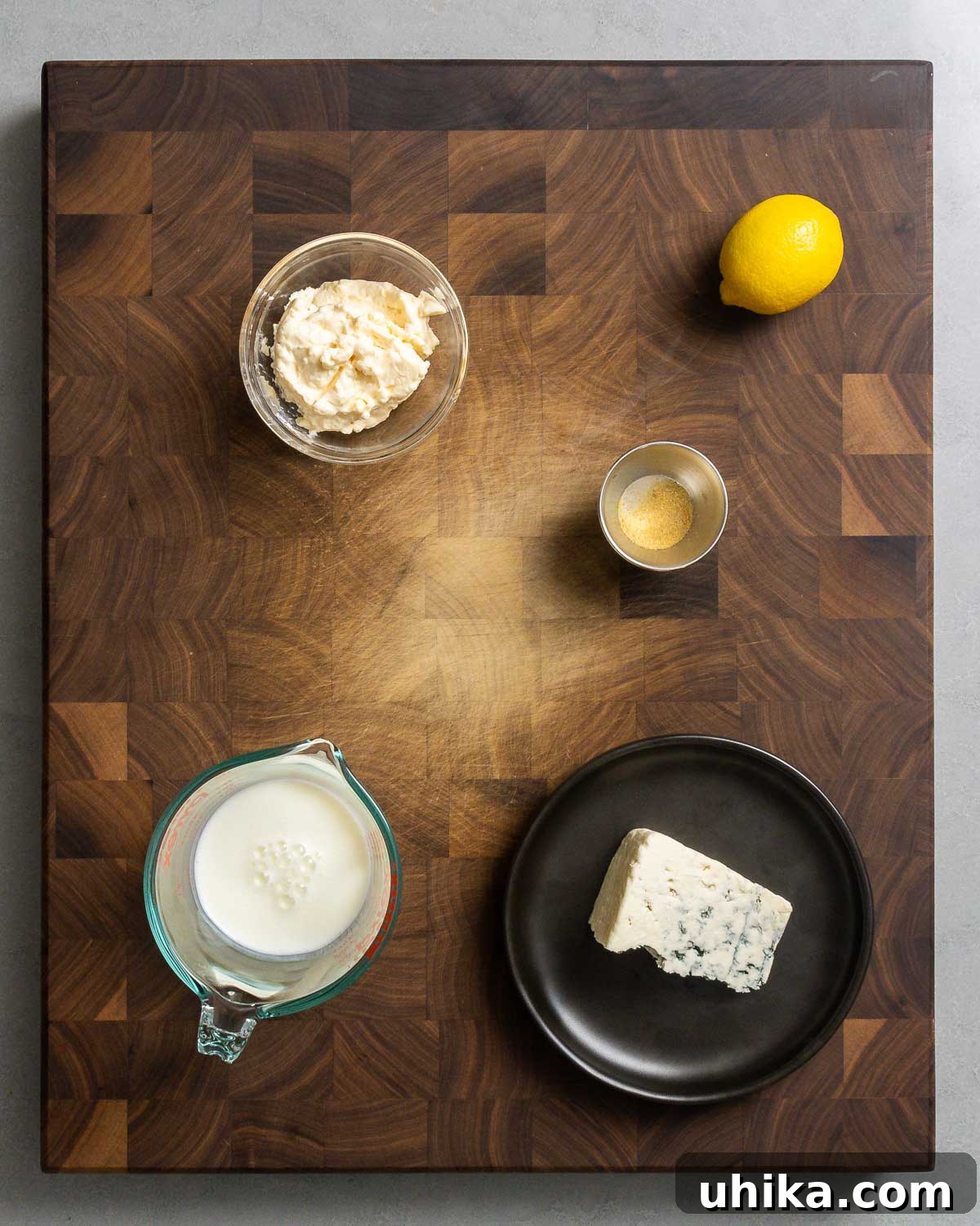 A selection of ingredients for blue cheese dressing displayed on a light surface: mayonnaise, fresh lemon, garlic powder, buttermilk, and a block of blue cheese.