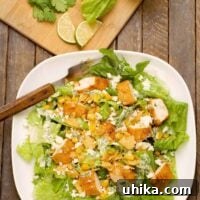An overhead view of a plate of Mexican-style Caesar salad; romaine lettuce, seasoned chicken tenders, corn, queso fresco, and tortilla chips, with a cutting board with fresh lime slices and cilantron in the background.