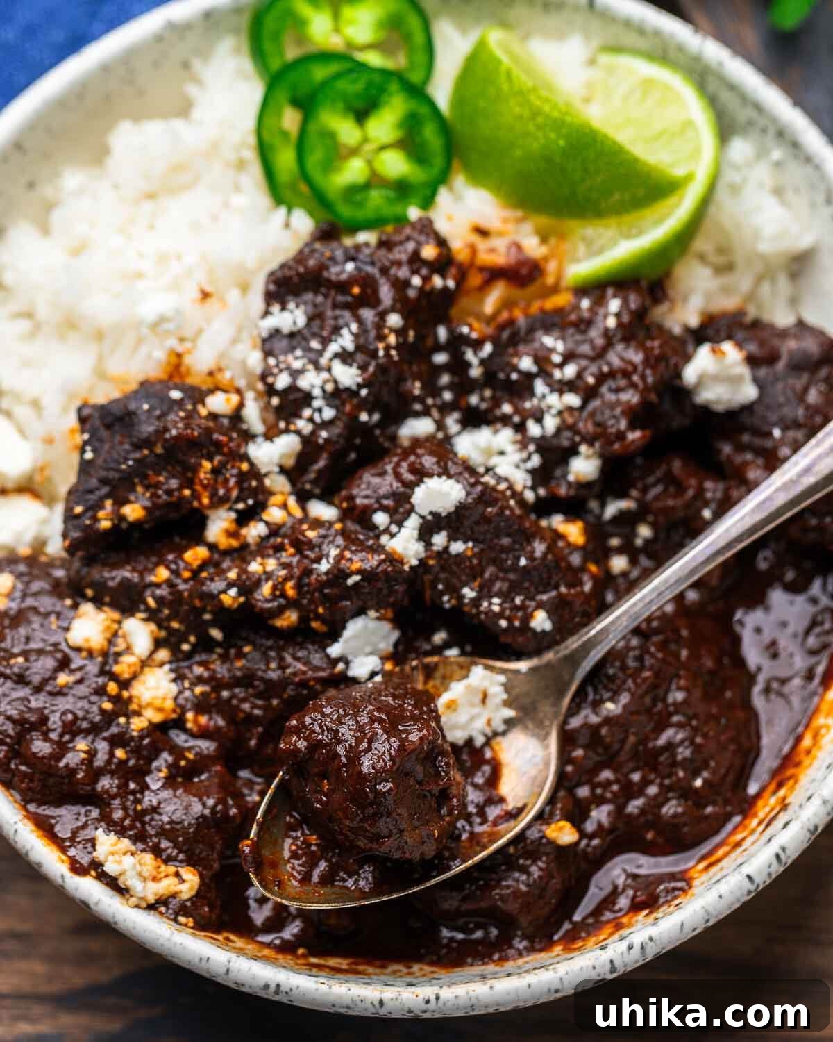 Crimson Beef Chili 2 White bowl with chili colorado, white rice, and garnished with limes, cilantro, and cotija cheese.