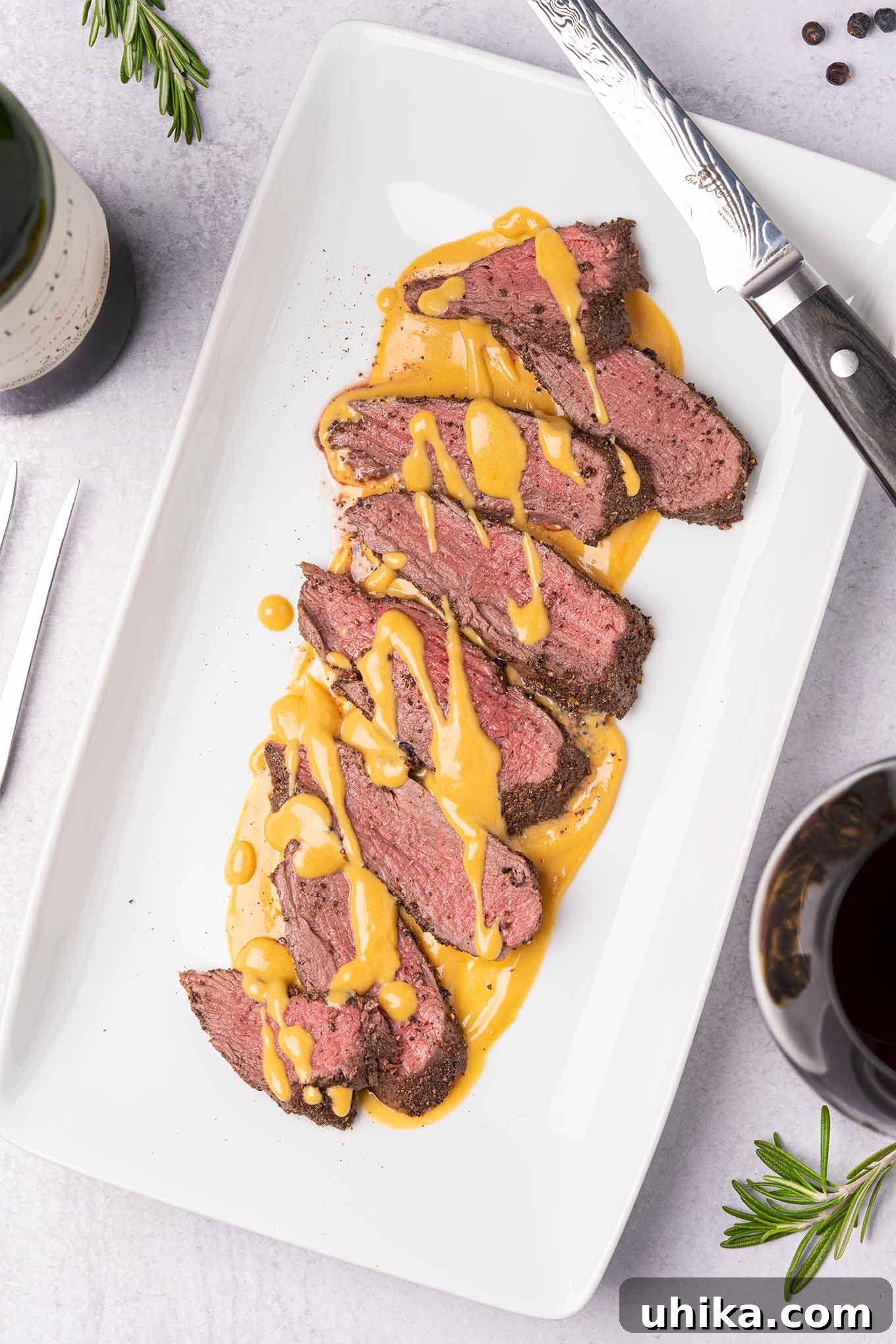 Juniper-Kissed Nordic Venison Tenderloin 2 An overhead view of a white serving platter with slices of spice-crusted, roast venison tenderloin, drizzled with gjetost cheese sauce, a carving knife on the edge of the plate, and sprigs of fresh rosemary, peppercorns, and a glass of red wine off to the side.