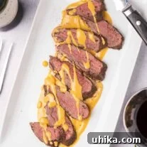 Juniper-Kissed Nordic Venison Tenderloin 4 An overhead view of a white serving platter with slices of spice-crusted, roast venison tenderloin, drizzled with gjetost cheese sauce, a carving knife on the edge of the plate, and sprigs of fresh rosemary, peppercorns, and a glass of red wine off to the side.