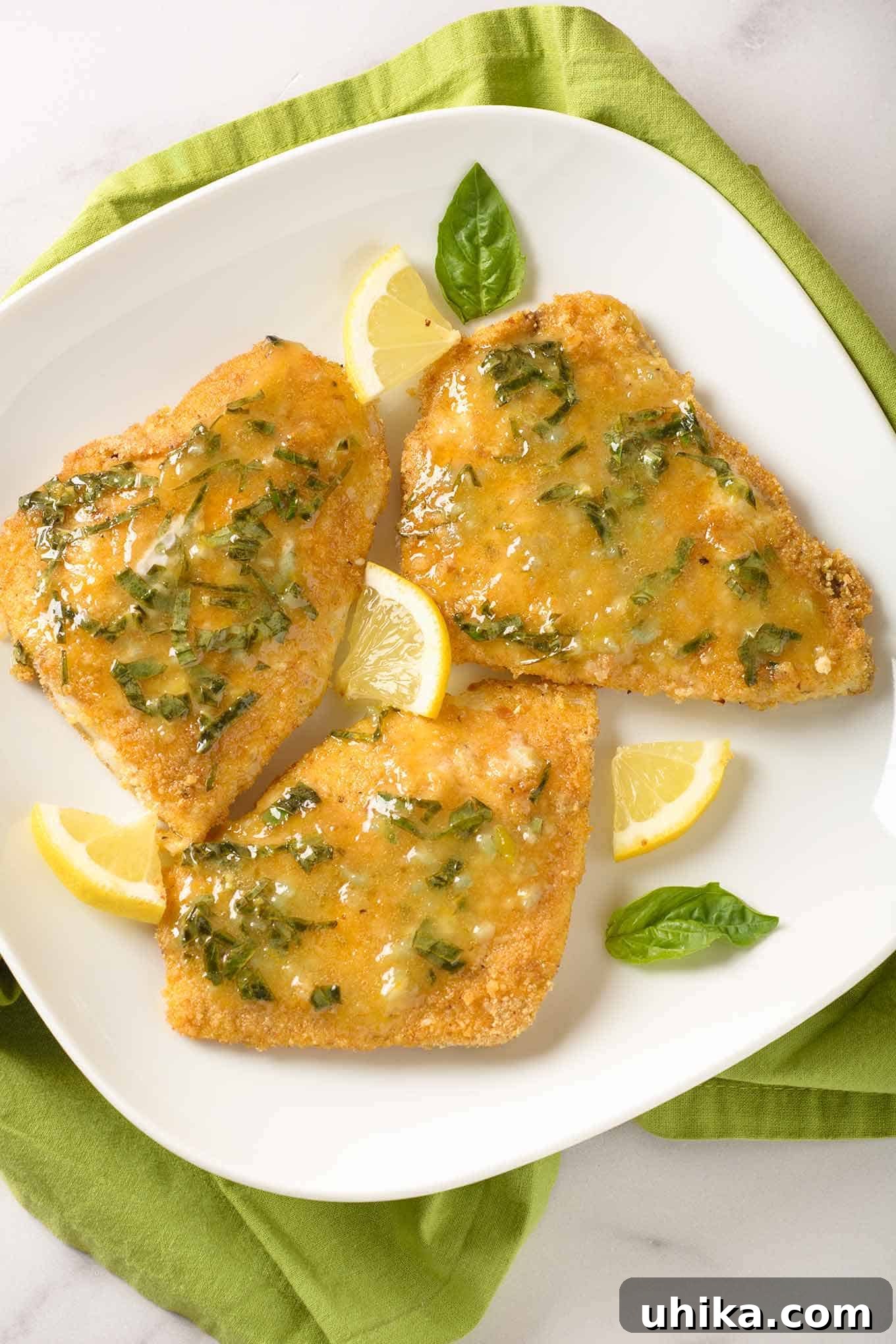 Golden Oven-Crisped Fish with Lemon Basil Butter 2 Three pieces of breaded, oven-fried fish fillets topped with lemon butter infused with fresh basil and garnished with basil leaves and lemon wedges, ready to be served.