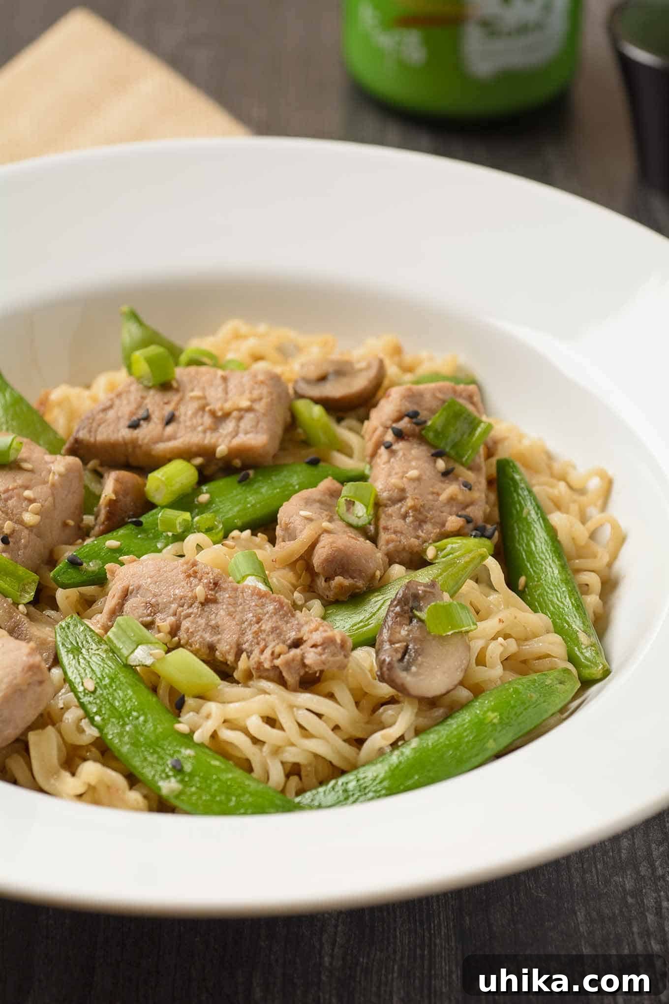Skillet Pork Ramen 3 A shallow bowl of ramen noodles tossed with stir-fried pork, sugar snap peas, and scallions with a bottle of sake in the background, highlighting an easy one-pan dinner.