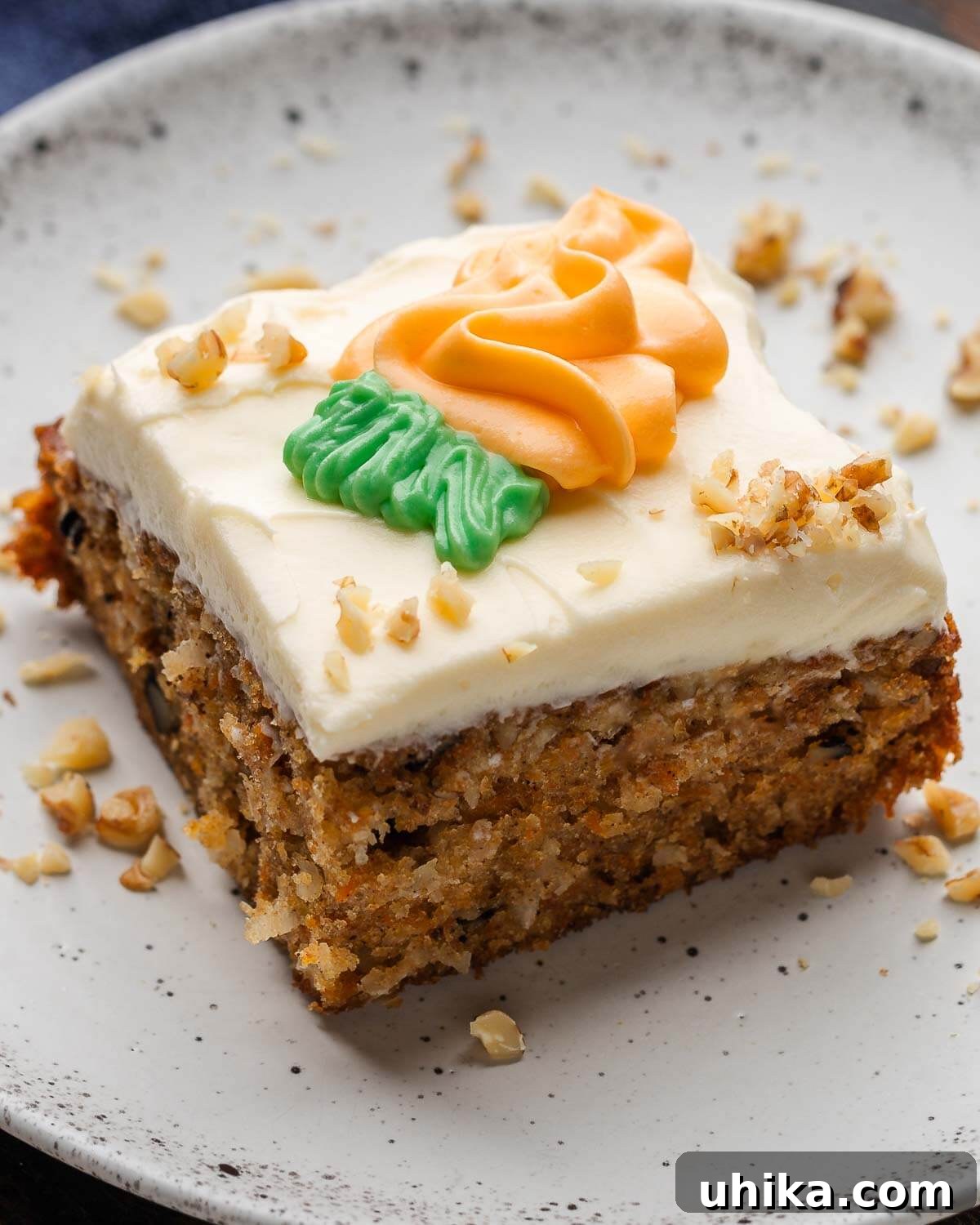 Carrot Cake Bliss 2 A generous slice of moist carrot cake topped with creamy white cream cheese frosting, garnished with finely chopped walnuts, presented on a white plate with walnuts sprinkled around the edges.
