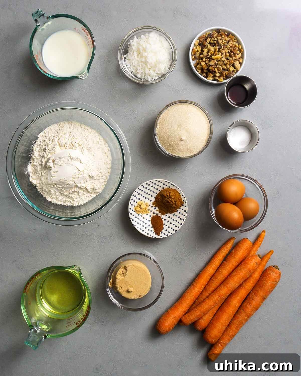 Carrot Cake Bliss 3 All ingredients for the carrot cake laid out on a white surface, including milk, coconut flakes, walnuts, vanilla extract, baking soda, all-purpose flour, granulated sugar, ground cinnamon, nutmeg, ginger, eggs, vegetable oil, brown sugar, and a pile of fresh carrots.