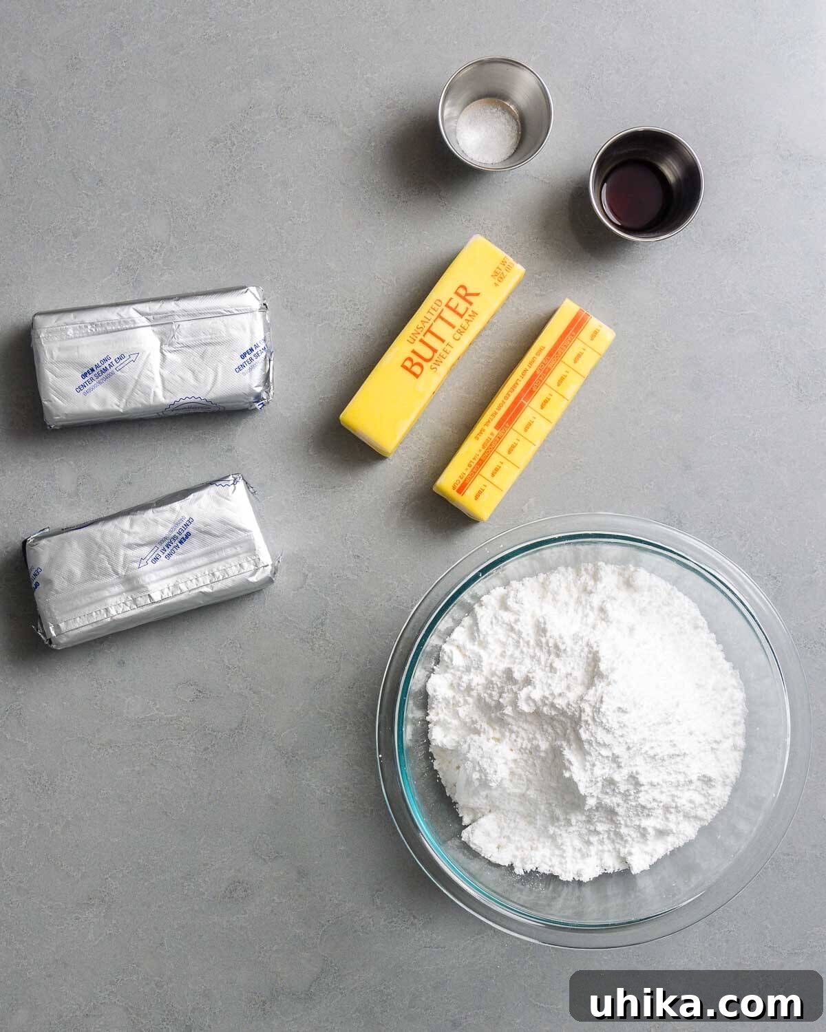 Carrot Cake Bliss 4 Ingredients for cream cheese frosting displayed on a white surface: salt, vanilla extract, blocks of cream cheese, unsalted butter, and powdered sugar.