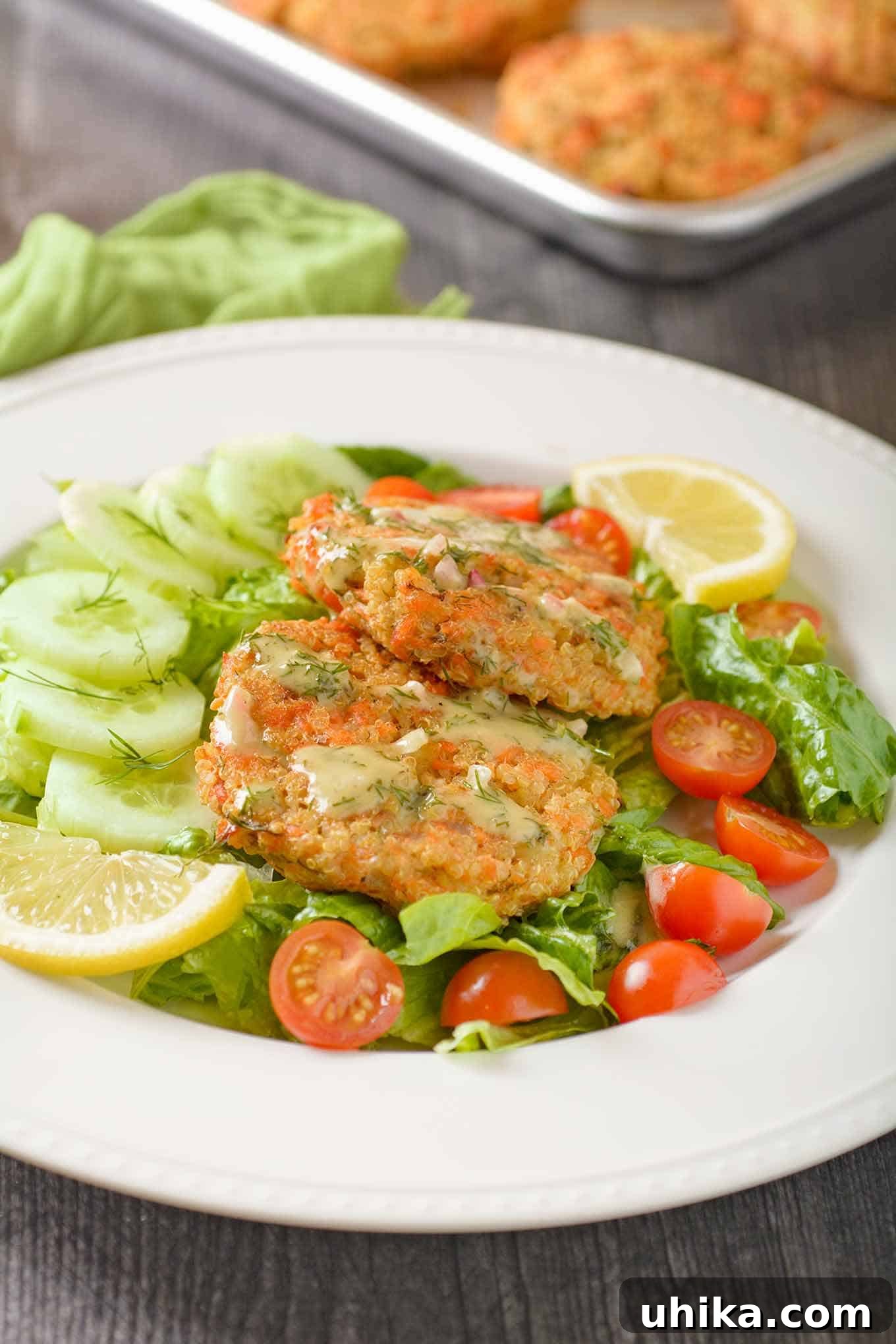 Savory Salmon Quinoa Cakes 3 A close-up serving of baked salmon quinoa cakes plated with a fresh green salad, accompanied by creamy lemon and dill dressing. In the blurred background, a sheet pan with more salmon patties hints at a delicious batch in progress.