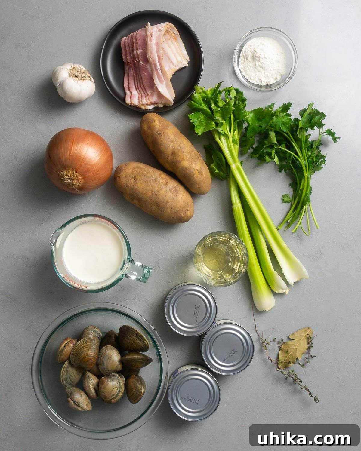 Authentic New England Clam Chowder 3 A flat lay of all ingredients for New England clam chowder: fresh garlic, crisp bacon, all-purpose flour, a whole onion, russet potatoes, celery stalks, fresh parsley, heavy cream, white wine, canned chopped clams, fresh littleneck clams, sprigs of thyme, and bay leaves.