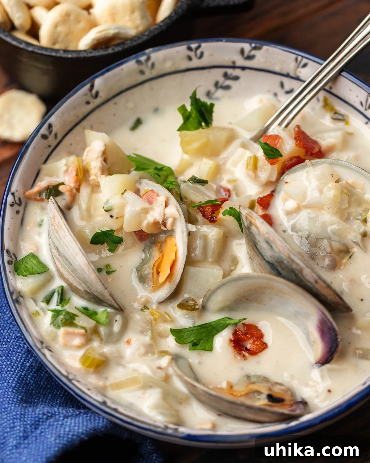 Authentic New England Clam Chowder 6 A smaller bowl of New England clam chowder, accompanied by oyster crackers in the background, presented on a rustic wooden surface.