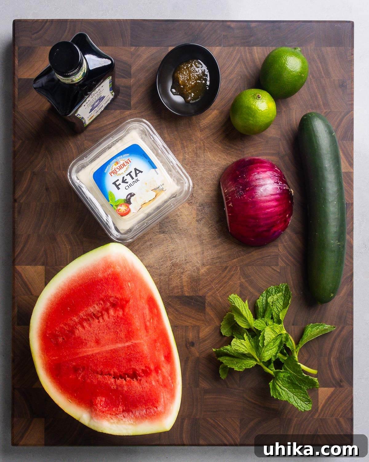 Refreshing Watermelon Feta Cucumber Mint Salad 3 A flat lay shot of fresh ingredients laid out on a clean surface. The items include a bottle of extra virgin olive oil, a small jar of honey, whole fresh limes, a block of creamy feta cheese, thinly sliced red onion, crisp cucumber, vibrant red cubed watermelon, and a bunch of fresh mint leaves. All components for the salad are clearly visible and ready for preparation.