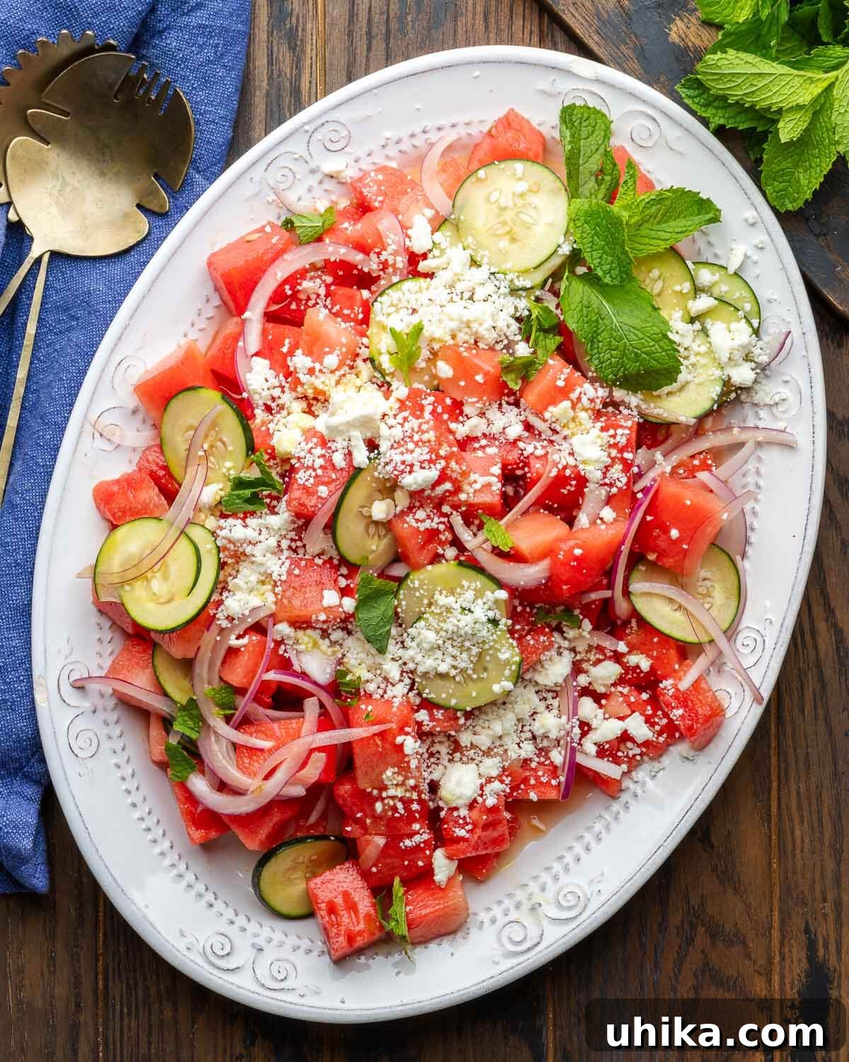 Refreshing Watermelon Feta Cucumber Mint Salad 4 A large, elegant white platter generously piled with the vibrant Watermelon Feta Salad, beautifully garnished with fresh mint sprigs. The platter rests on a rustic wooden table, flanked by decorative bronze serving spoons and a small cutting board holding extra mint. The scene evokes a feeling of effortless summer dining.