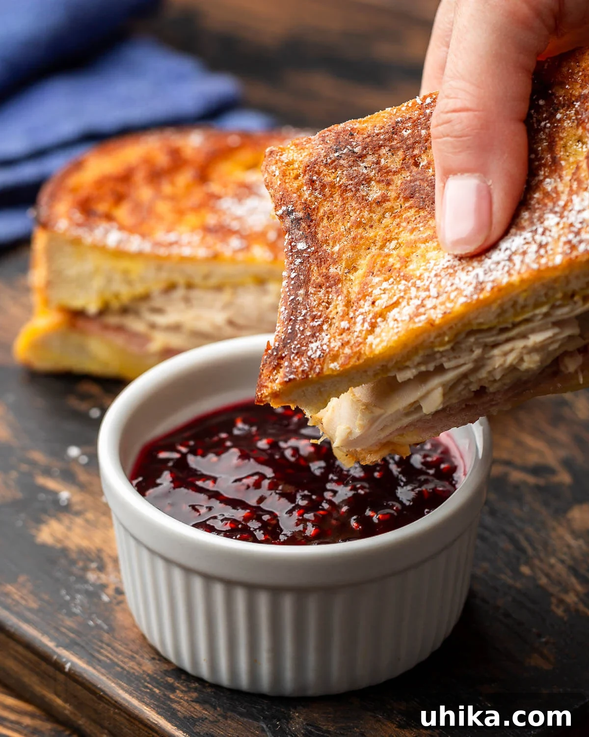 Monte Cristo's Reckoning 2 A perfectly golden-brown Monte Cristo sandwich being dipped into a small bowl of vibrant raspberry jam, highlighting its sweet and savory appeal.